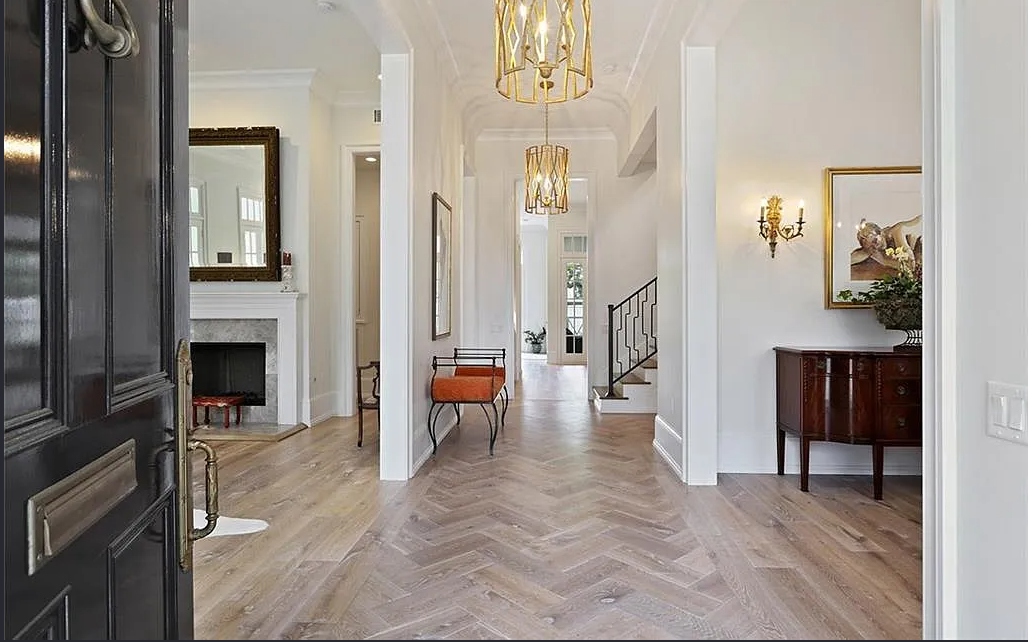 Elegant foyer with light-colored wooden floors, white walls, and classic decor including a black front door, gold chandeliers, a fireplace with a large mirror, and a small orange bench.