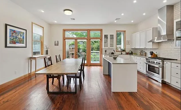 Bright, modern kitchen with white cabinets, stainless steel stove, and a large wooden dining table with six chairs, large windows and sliding door leading outside with greenery.
