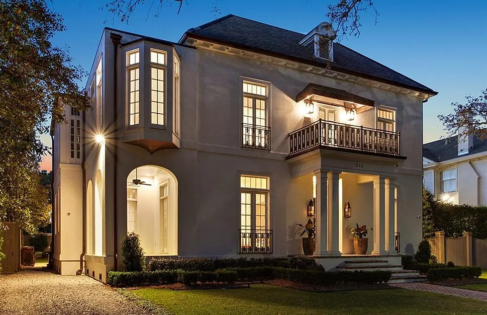 A large, two-story house illuminated at dusk, featuring a white exterior, tall bay windows, a balcony on the second floor, and a covered entrance with pillars.
