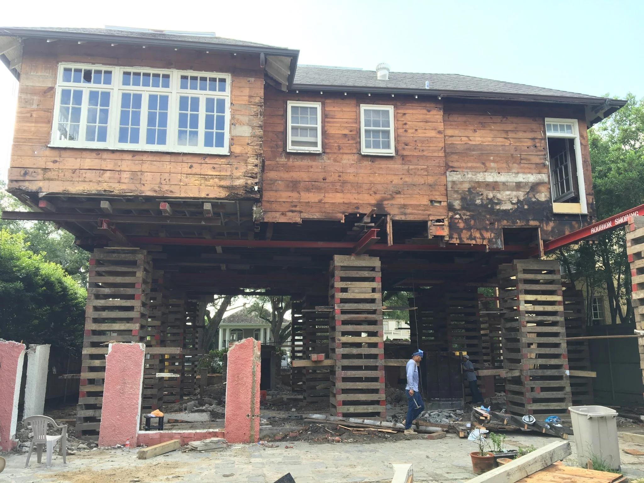 A house under construction, elevated on wooden stilts, with workers at the site and trees in the background.