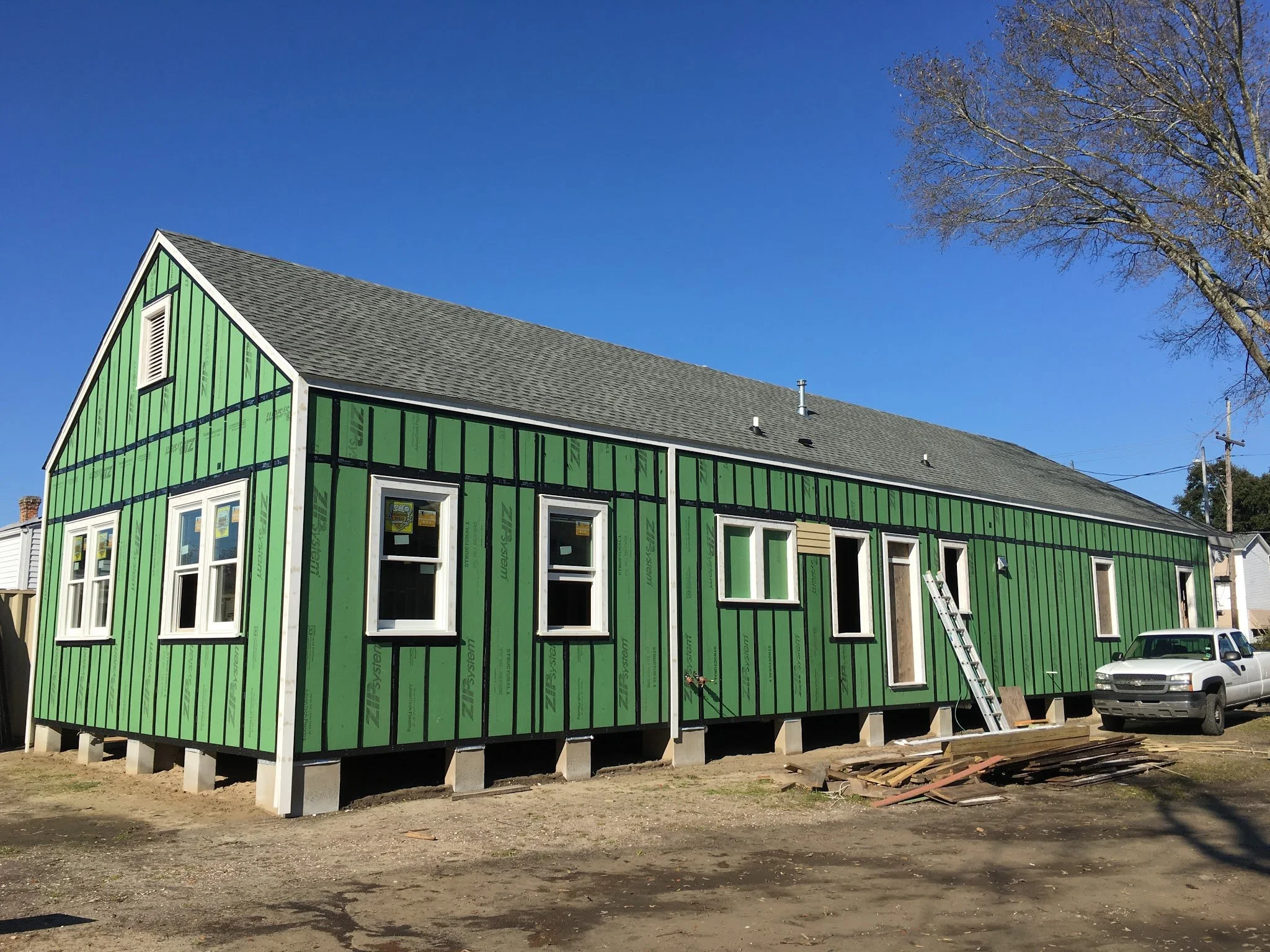 A house under construction with green sheathing, elevated on concrete blocks, with several windows installed, and surrounded by a lot with construction materials and a car parked nearby. The house has a gray shingle roof and is set against a clear bl