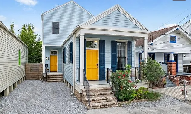 A two-story house painted white with blue shutters and a bright yellow front door, front porch with steps, surrounded by a small garden with bushes, and neighboring houses visible on each side.