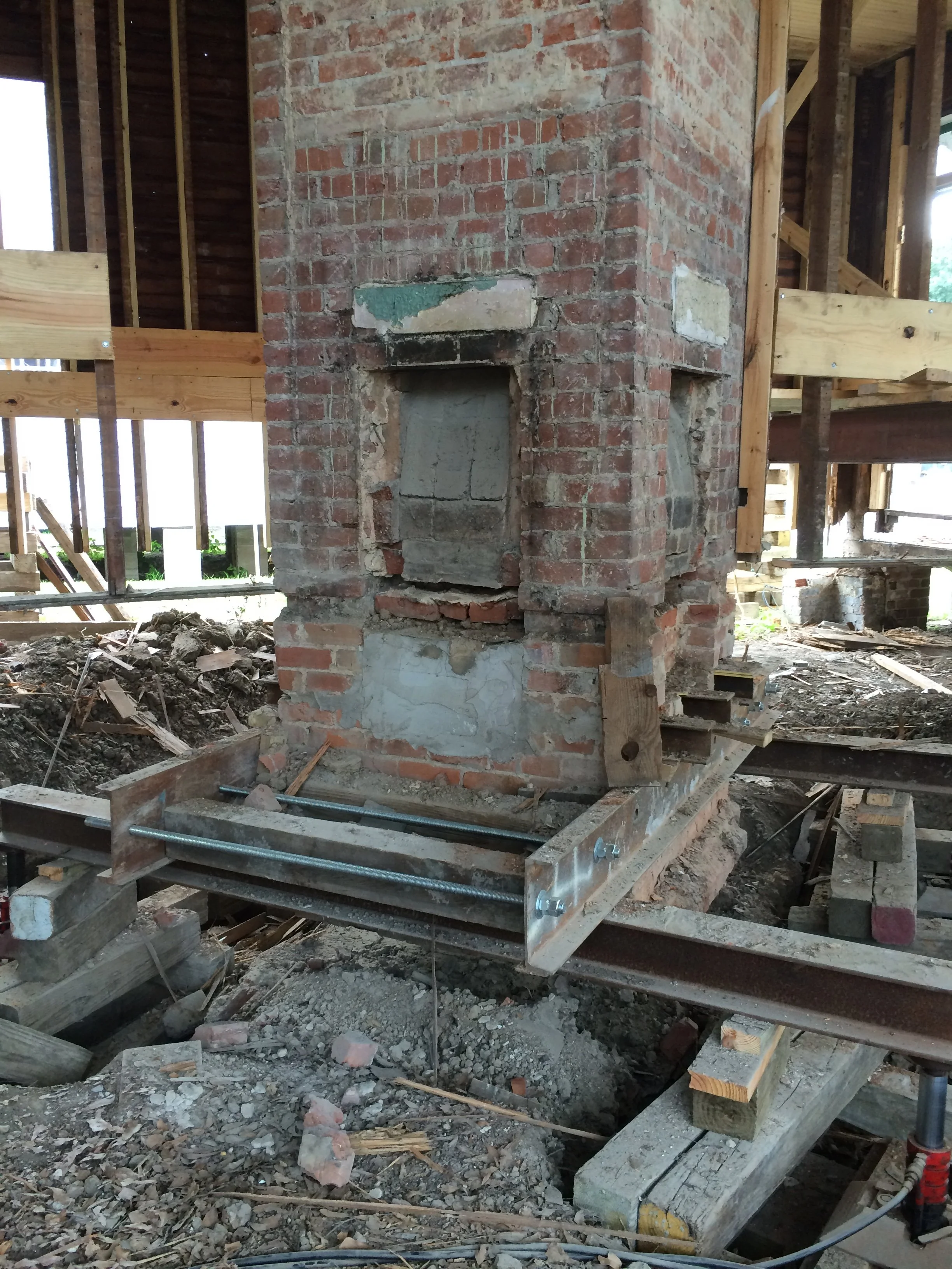 A brick chimney under construction inside a building at a construction site. There are wooden framing and scaffolding around it, with construction tools and debris on the ground.