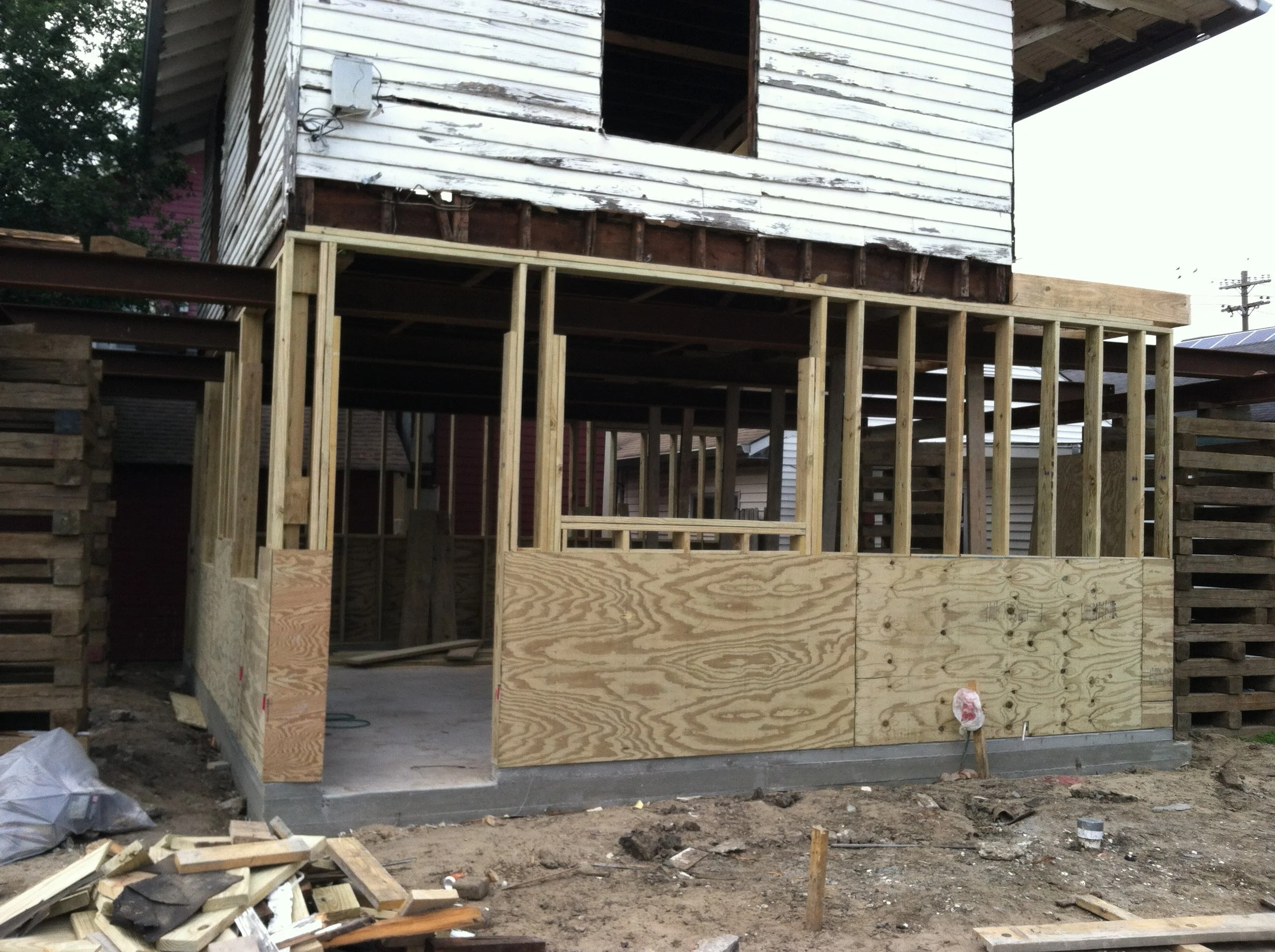 The image shows a house under construction with wooden framing. The house is raised on stilts, and the exterior walls are being installed, with some sections of the siding already in place while other parts are still open. There are construction mate