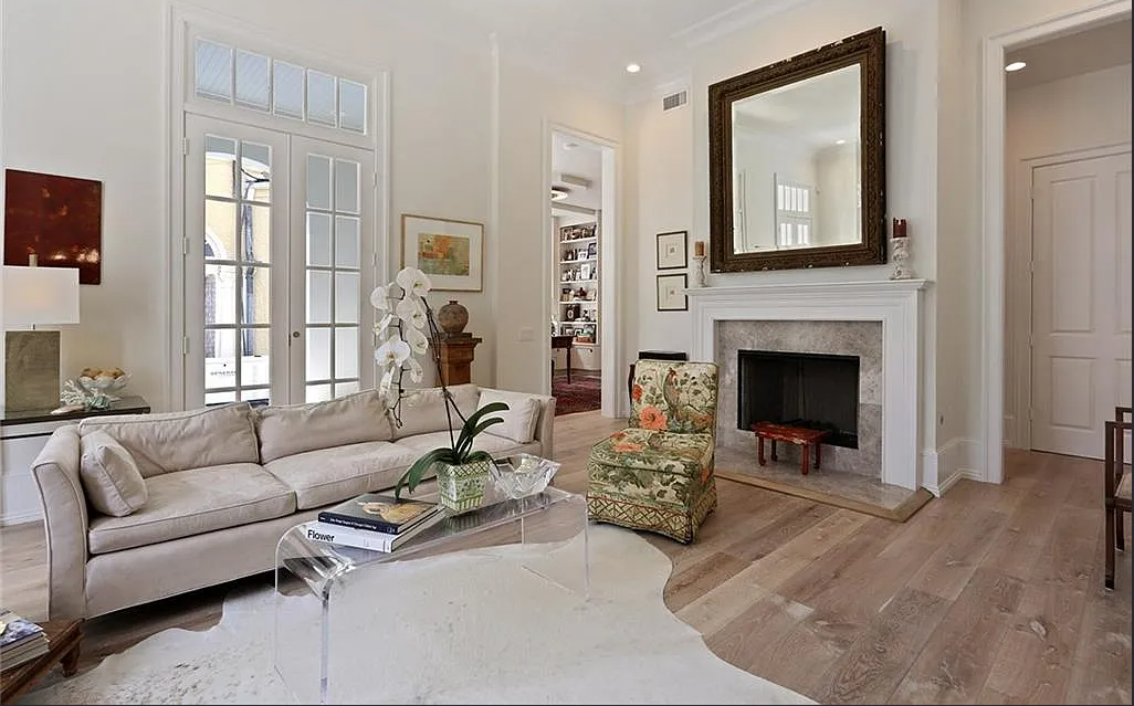 Living room with a beige sofa, glass coffee table, floral armchair, fireplace with a large mirror, and French doors allowing natural light.