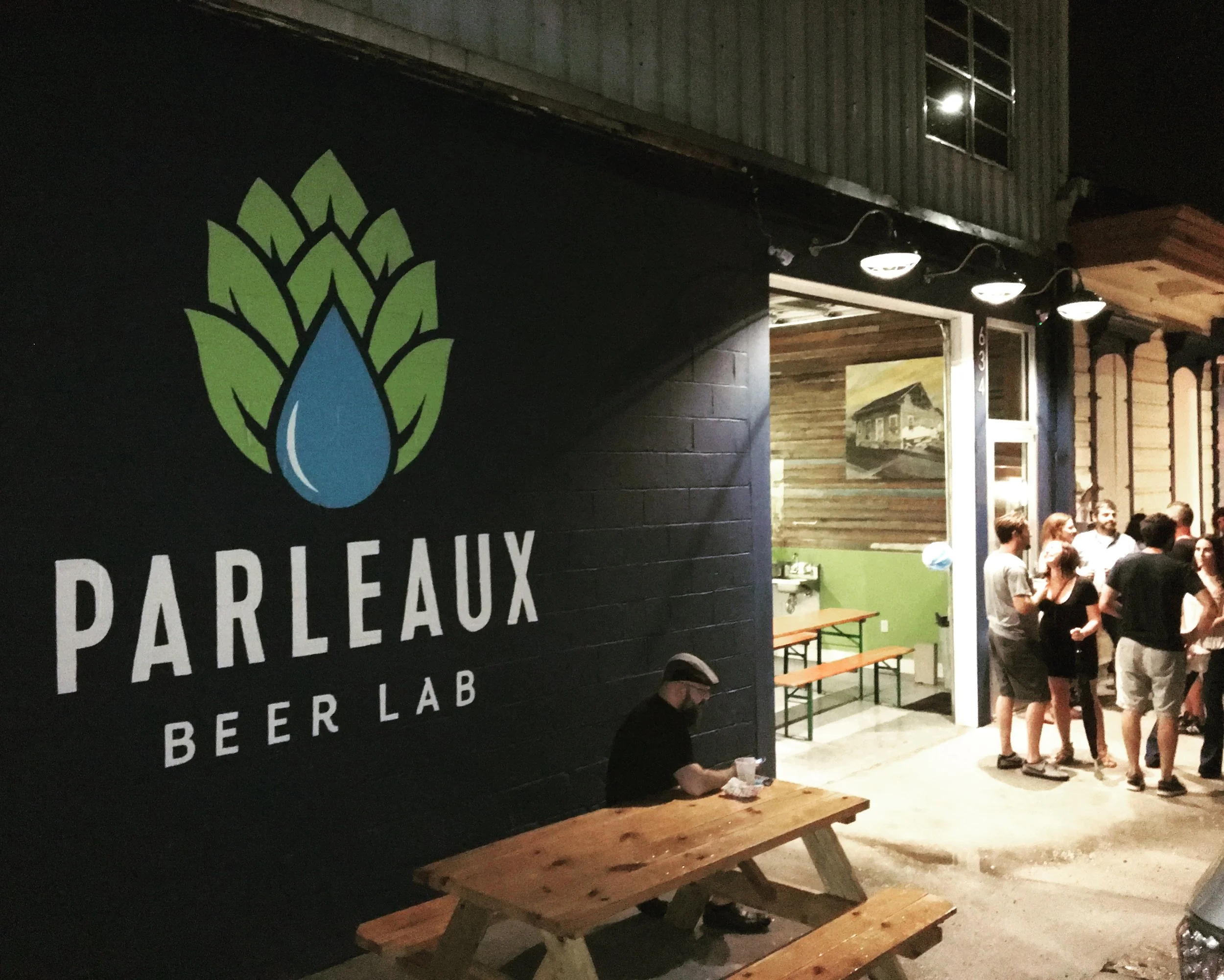 Night scene outside a brewery with a large black sign featuring a colorful hop cone with a blue water droplet in the middle and the partially visible words 'PARELAUX BREWERY LAB.' Inside, groups of people are gathered, some standing and talking, with