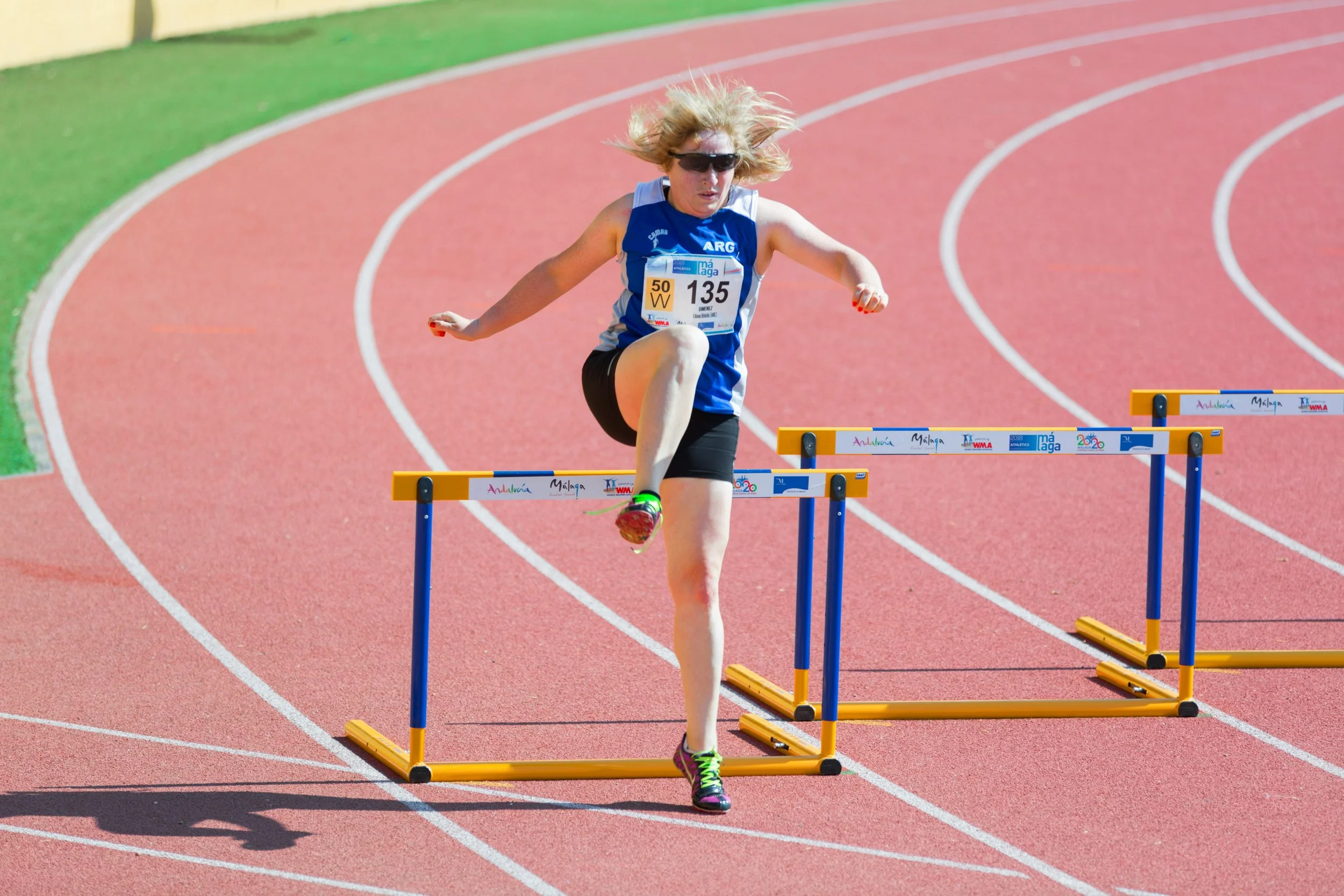 A female athlete with blonde hair wearing sunglasses, a blue and white sleeveless shirt, black shorts, and running shoes, participating in a hurdle race on a red track with green borders, mid-air clearing a hurdle.