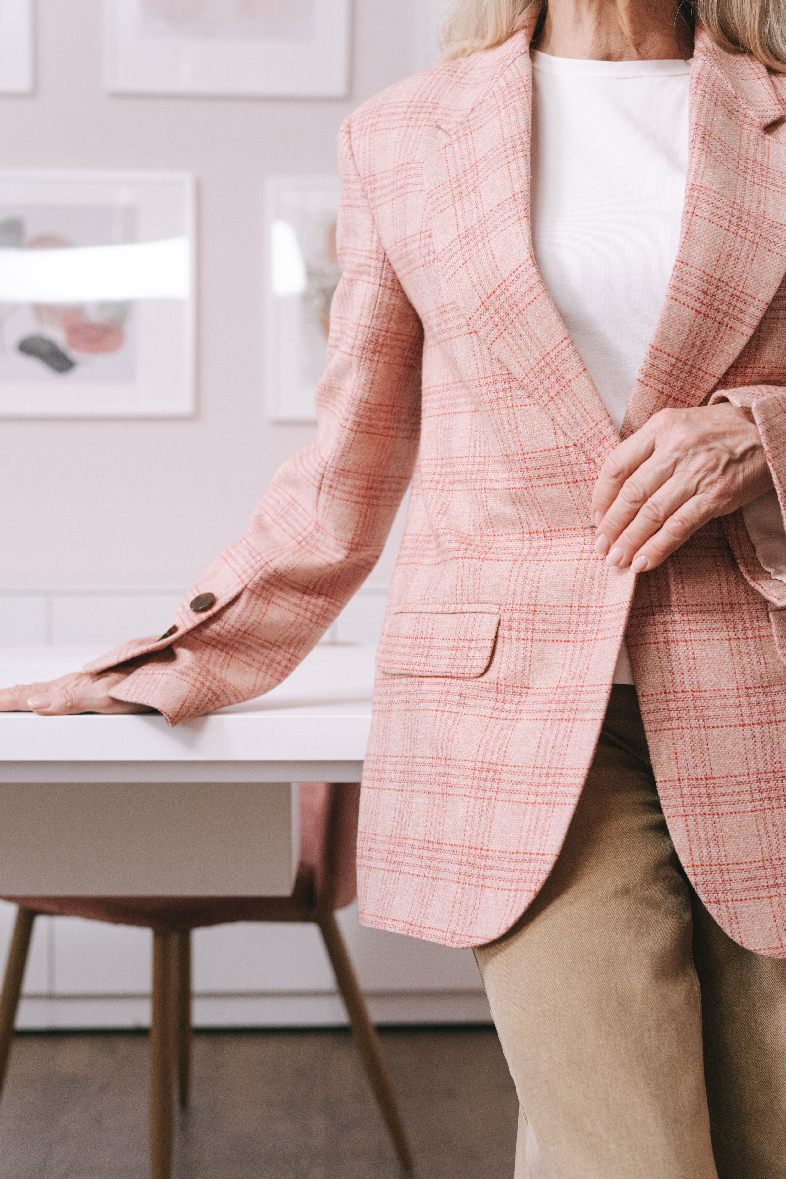 A woman wearing a pink plaid blazer, white top, and beige pants, standing in a modern, minimalistic room with art frames on the wall and a white desk.