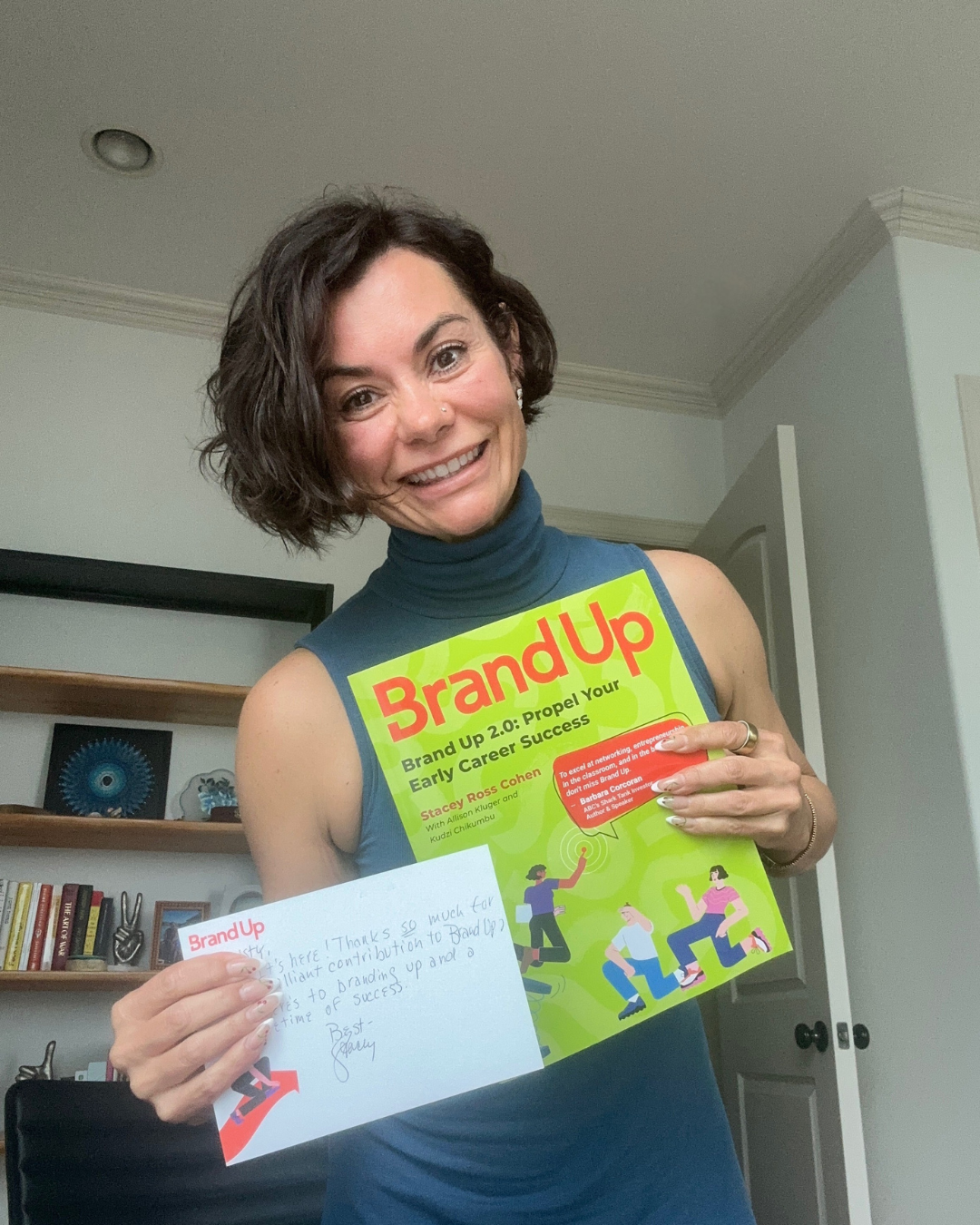 Woman with short dark hair smiling while holding a book titled 'Brand Up: Brand Up 2.0: Propel Your Early Career Success' in a room with bookshelves and decorative items.