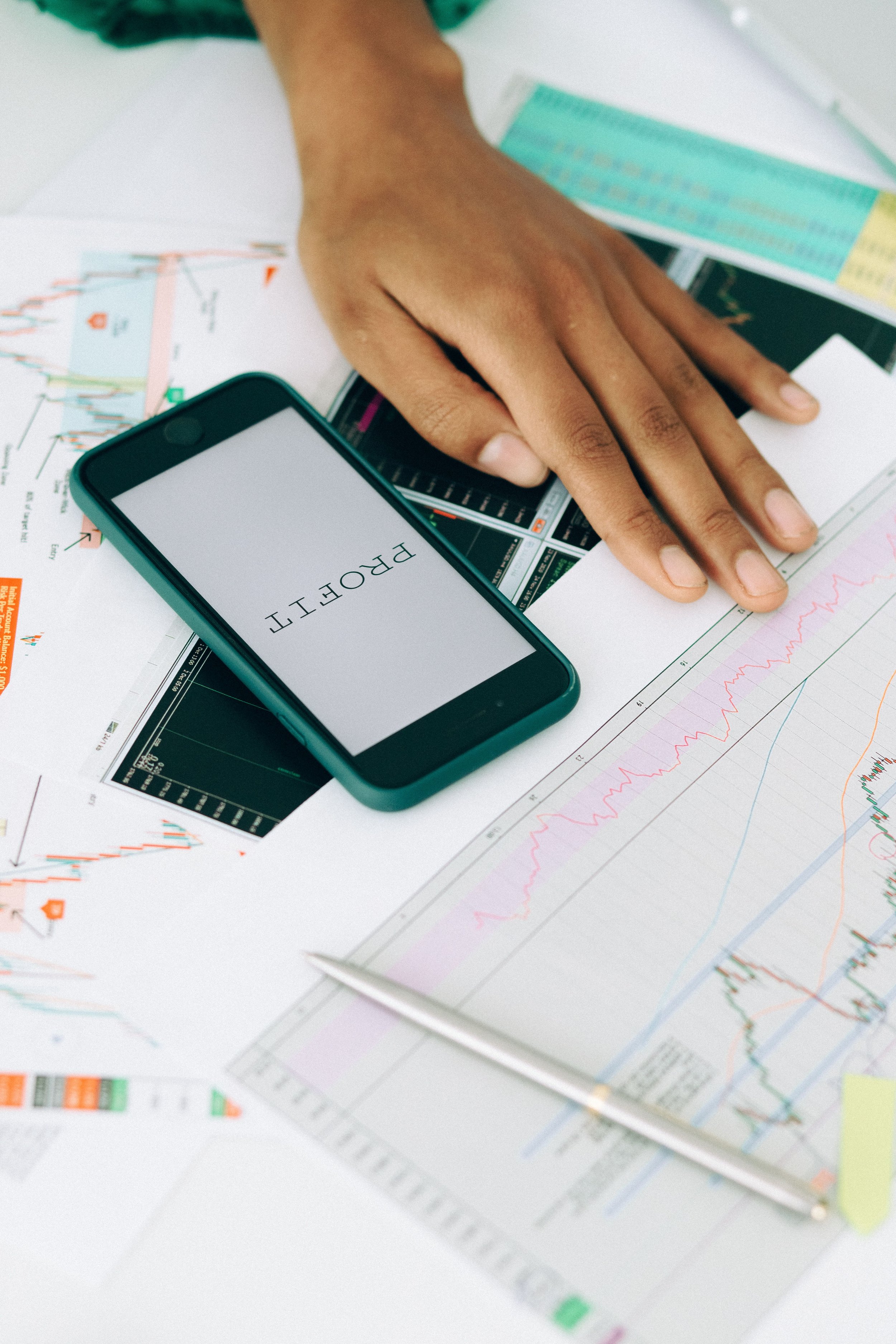 A hand resting on financial charts and graphs, with a smartphone displaying the word 'PROFIT' in uppercase letters.