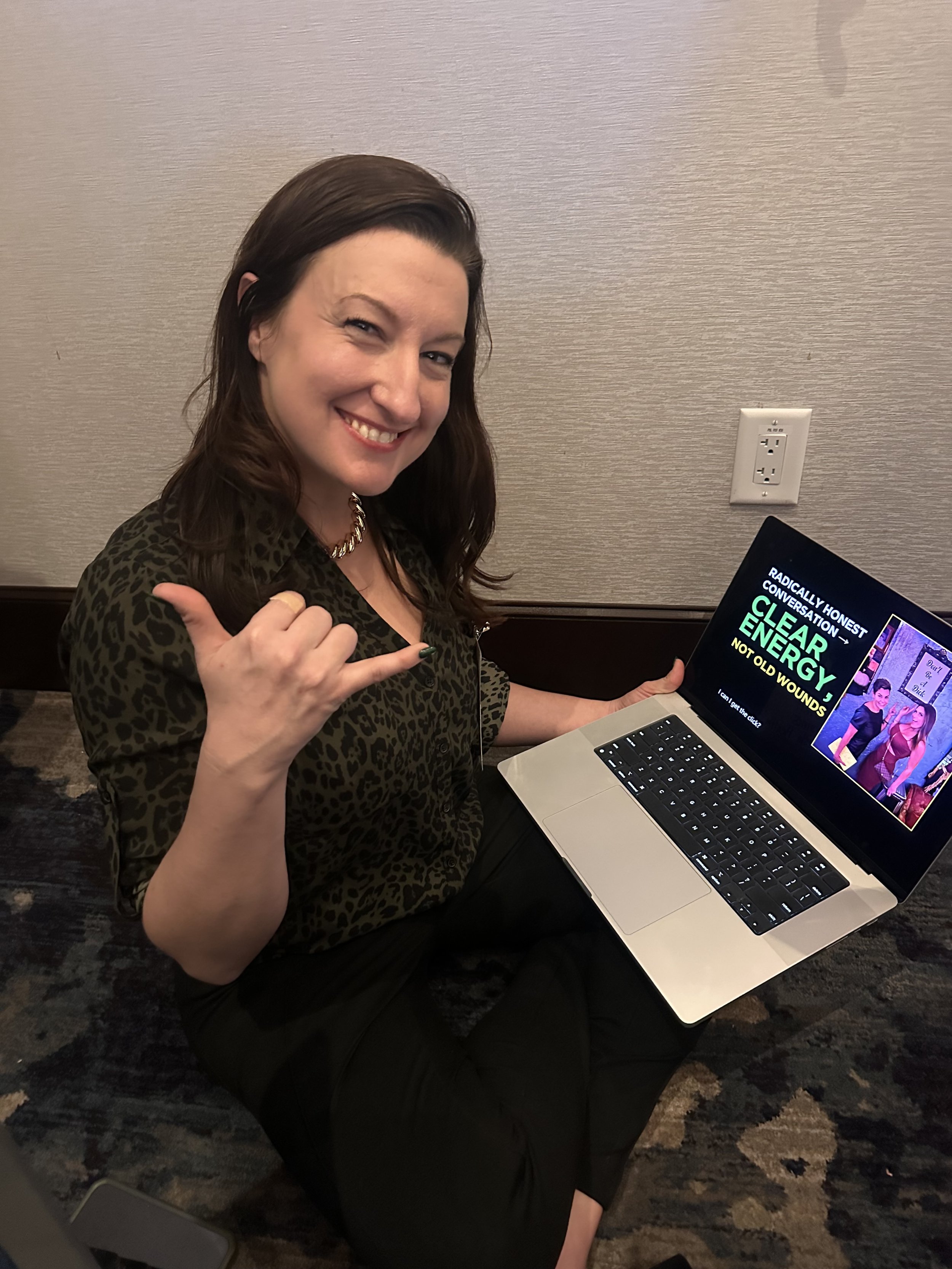 A smiling woman with dark brown hair, wearing a leopard print top and gold necklace, sitting on the floor with a laptop. She is making a shaka sign with her right hand, and the laptop screen displays a presentation titled 'Clear Energy' about energy and old wounds.