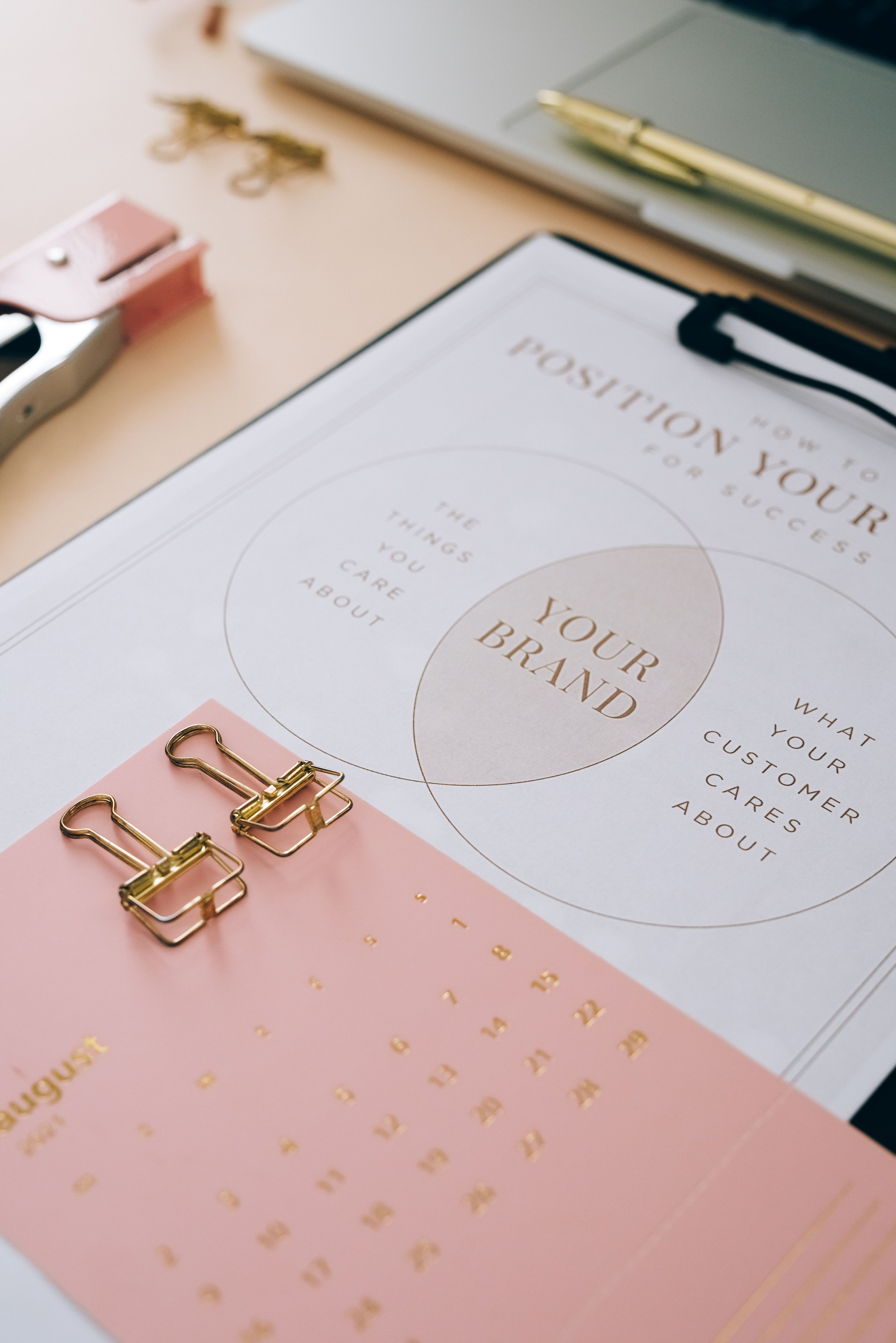 A workspace with various office supplies including a clipboard with a printed sheet titled 'How to Pose Your Logo & Show Success,' gold paper clips, a gold pen, and a calendar displaying the month of August with gold numbers and dates, all arranged on a light-colored desk.