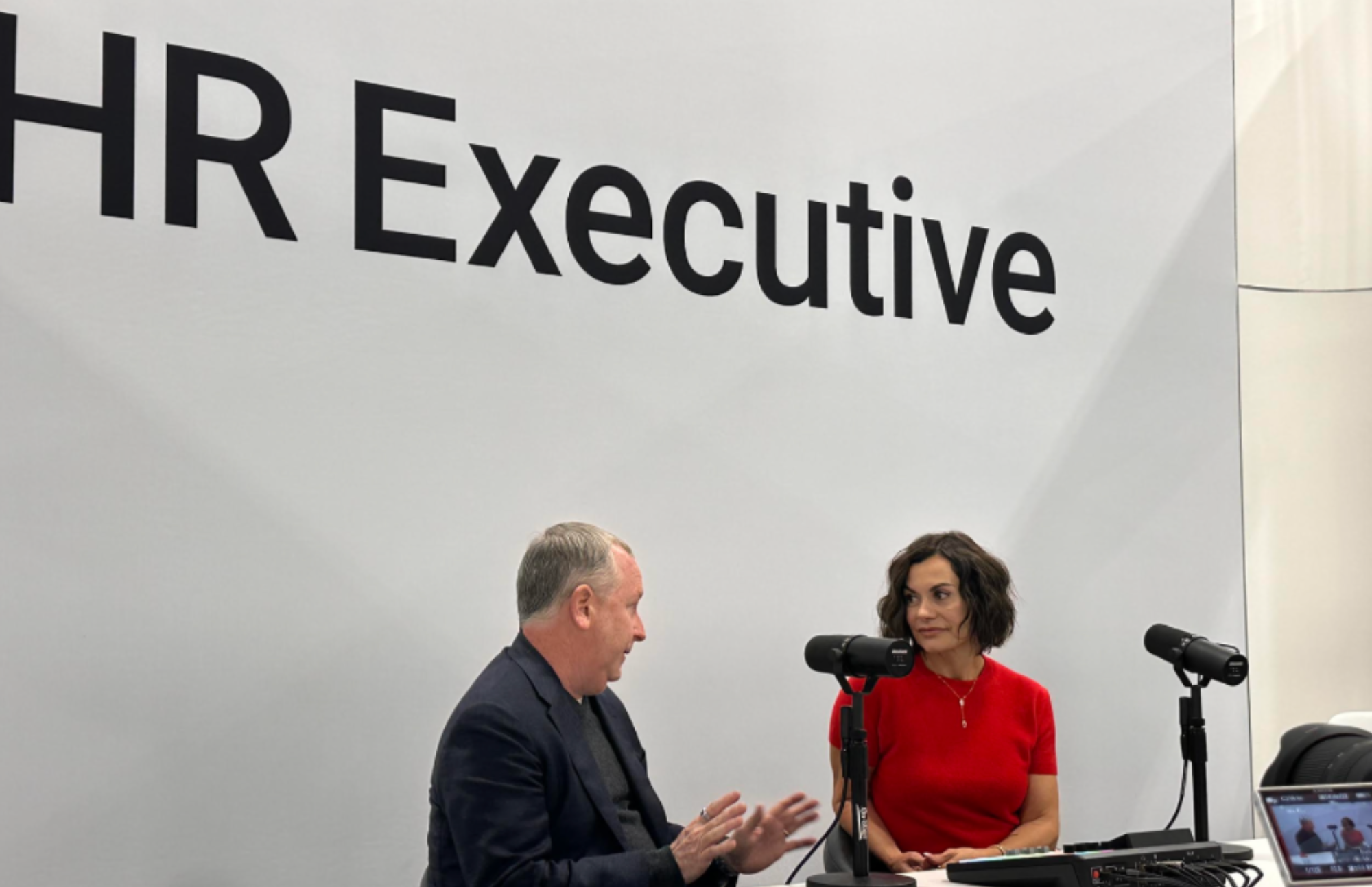 Two people, a man and a woman, sit at a table with microphones during a meeting or interview. The man has gray hair and is wearing a dark suit, while the woman has curly dark hair and is wearing a red top. A large sign behind them reads 'HR Executive.'
