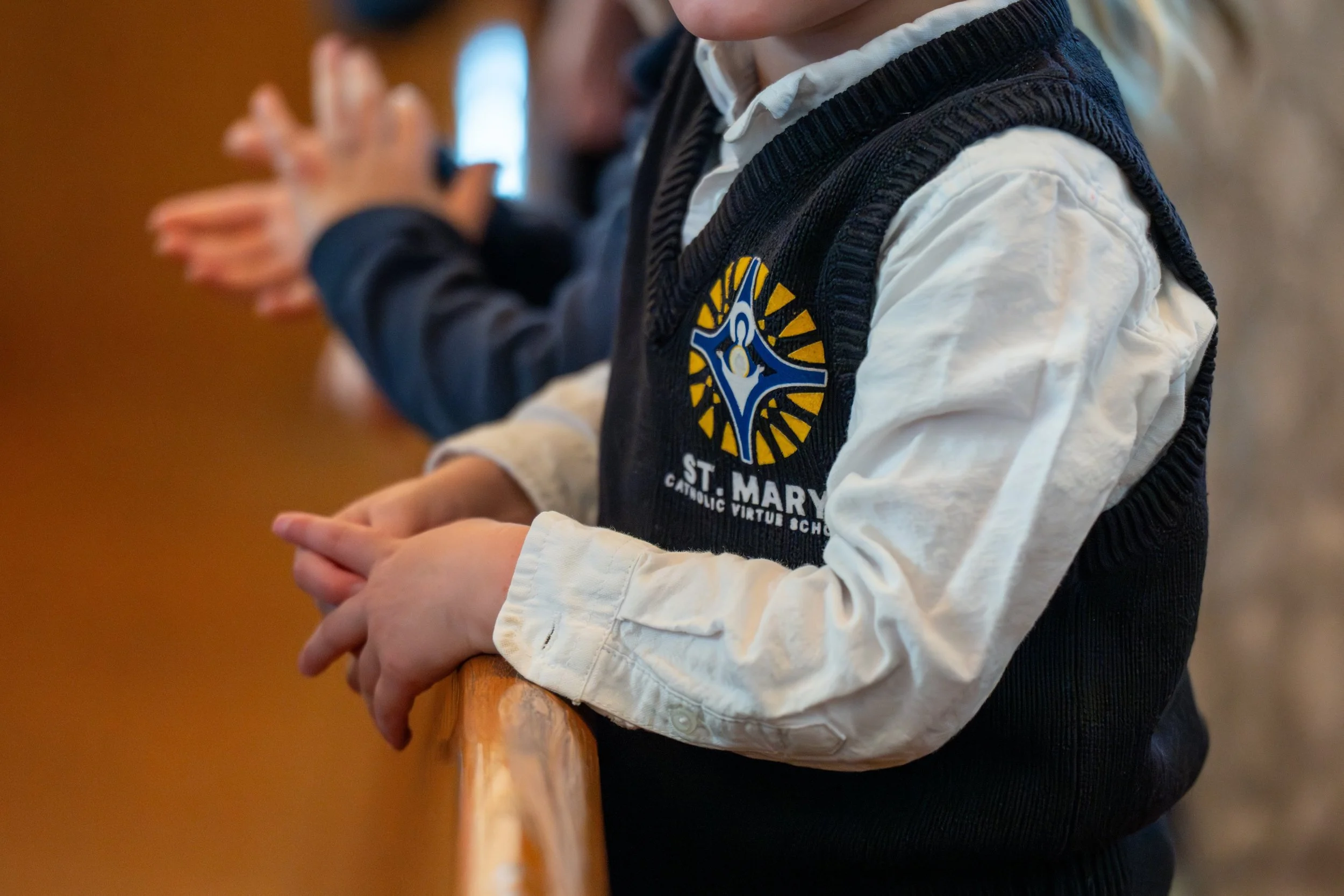 A child wearing a school uniform with a St. Mary logo is praying with hands clasped, kneeling at a wooden pew.