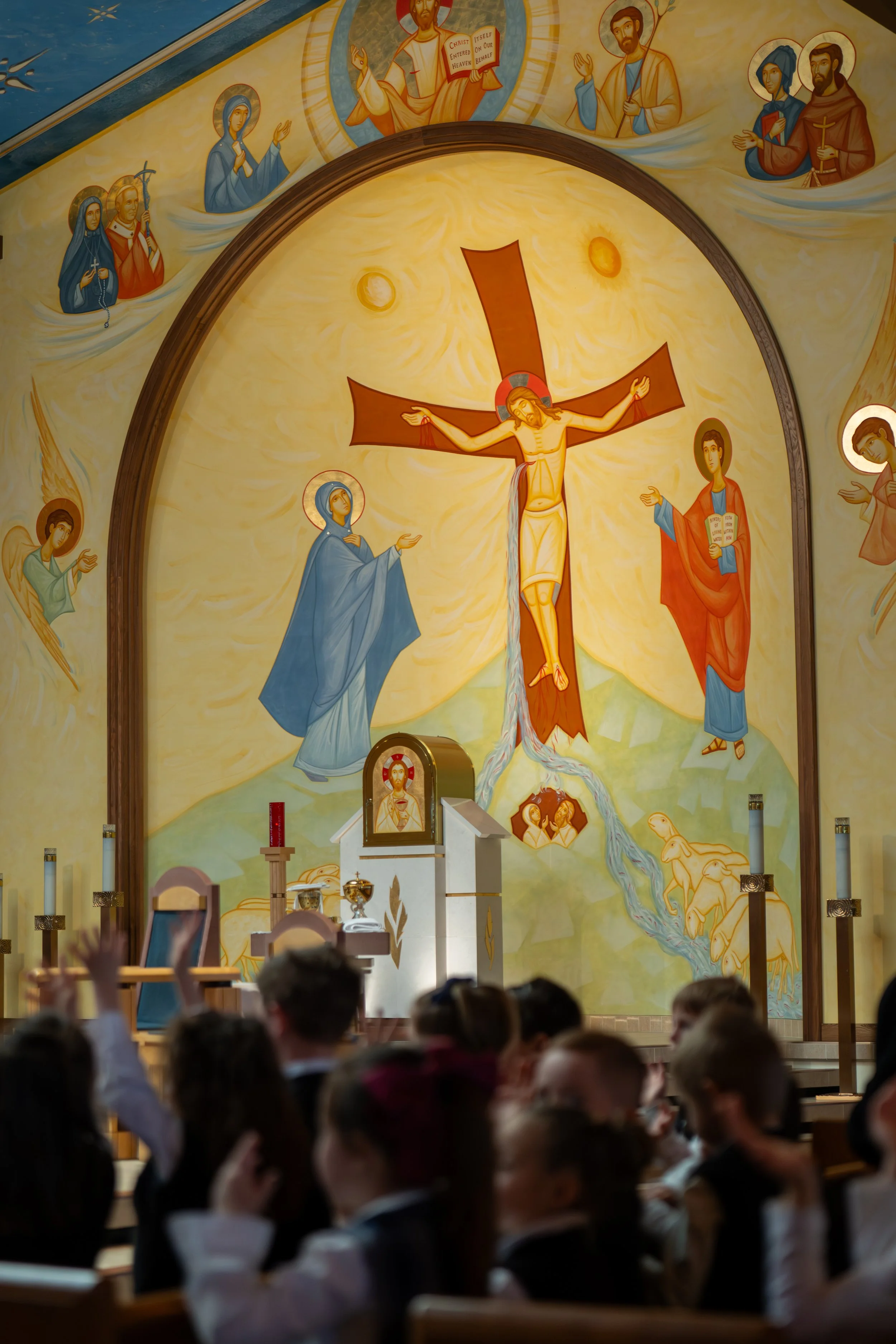 A Catholic church altar with a large religious mural depicting Jesus Christ on the cross, surrounded by various saints and religious figures, with congregation members sitting and praying in the foreground.