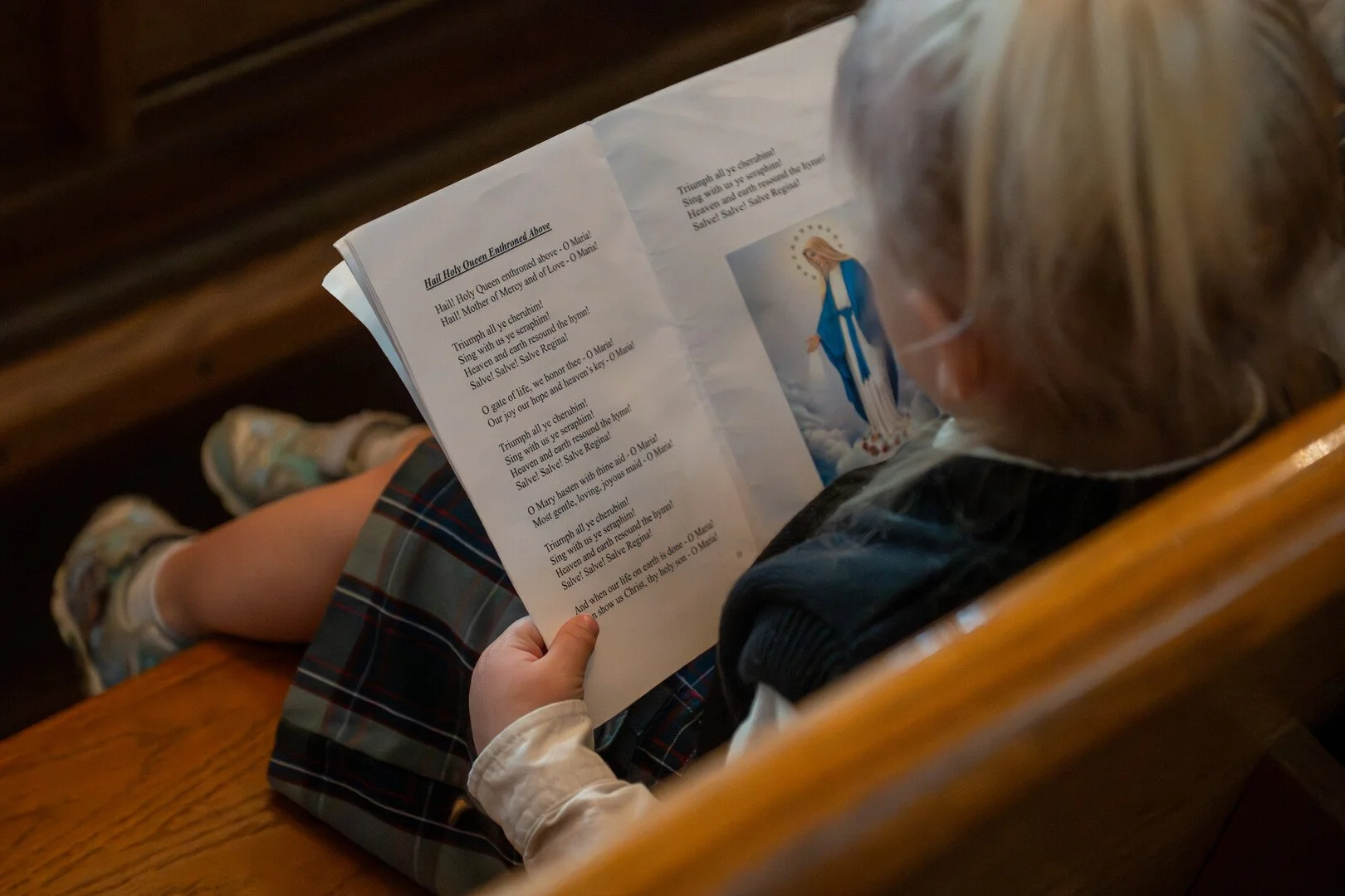A child sitting in a wooden pew reading a religious hymn book with an image of the Virgin Mary on the page.
