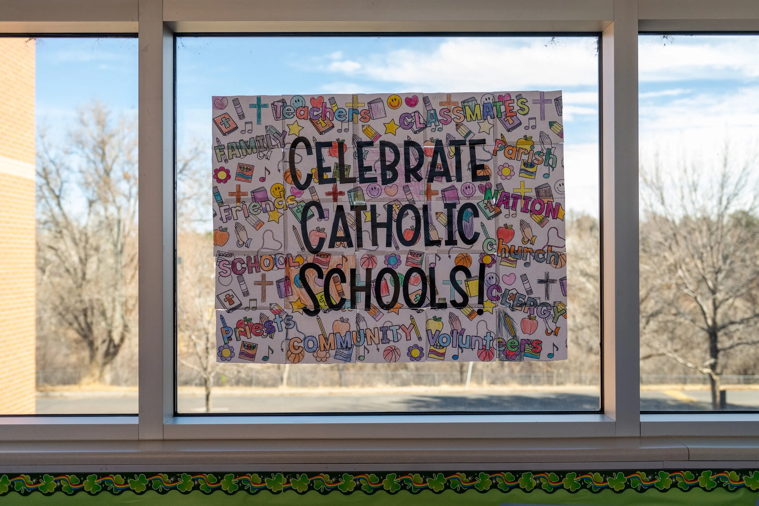 Colorful poster on window celebrating Catholic Schools Week with words like 'teachers,' 'classmates,' 'community,' and 'volunteers,' surrounded by school-themed illustrations such as pencils, books, crosses, and smiley faces, with a background view o