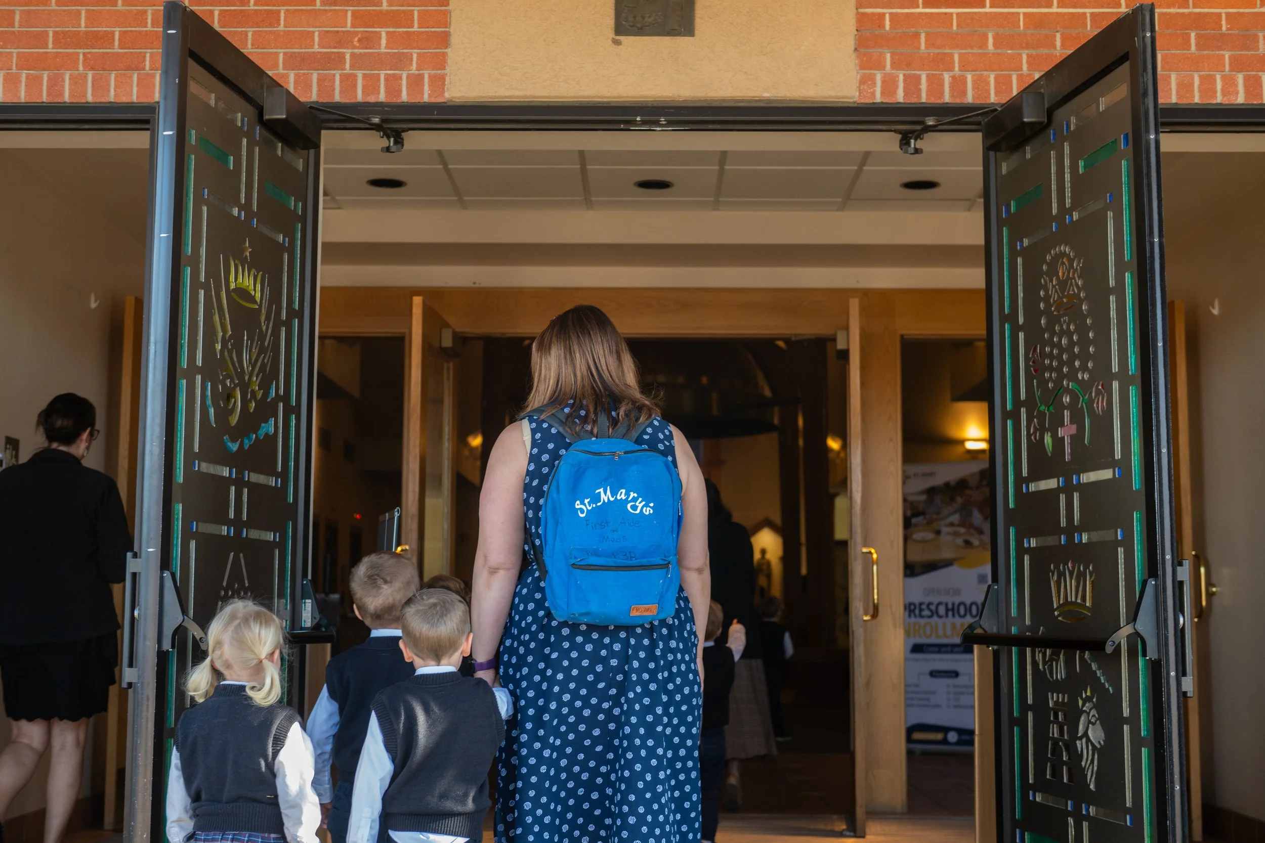 A group of children and a woman with a blue backpack standing at the entrance of a building, which looks like a school or preschool. The door is open, revealing an interior with signs of children’s activities and a sign that reads "Preschool".