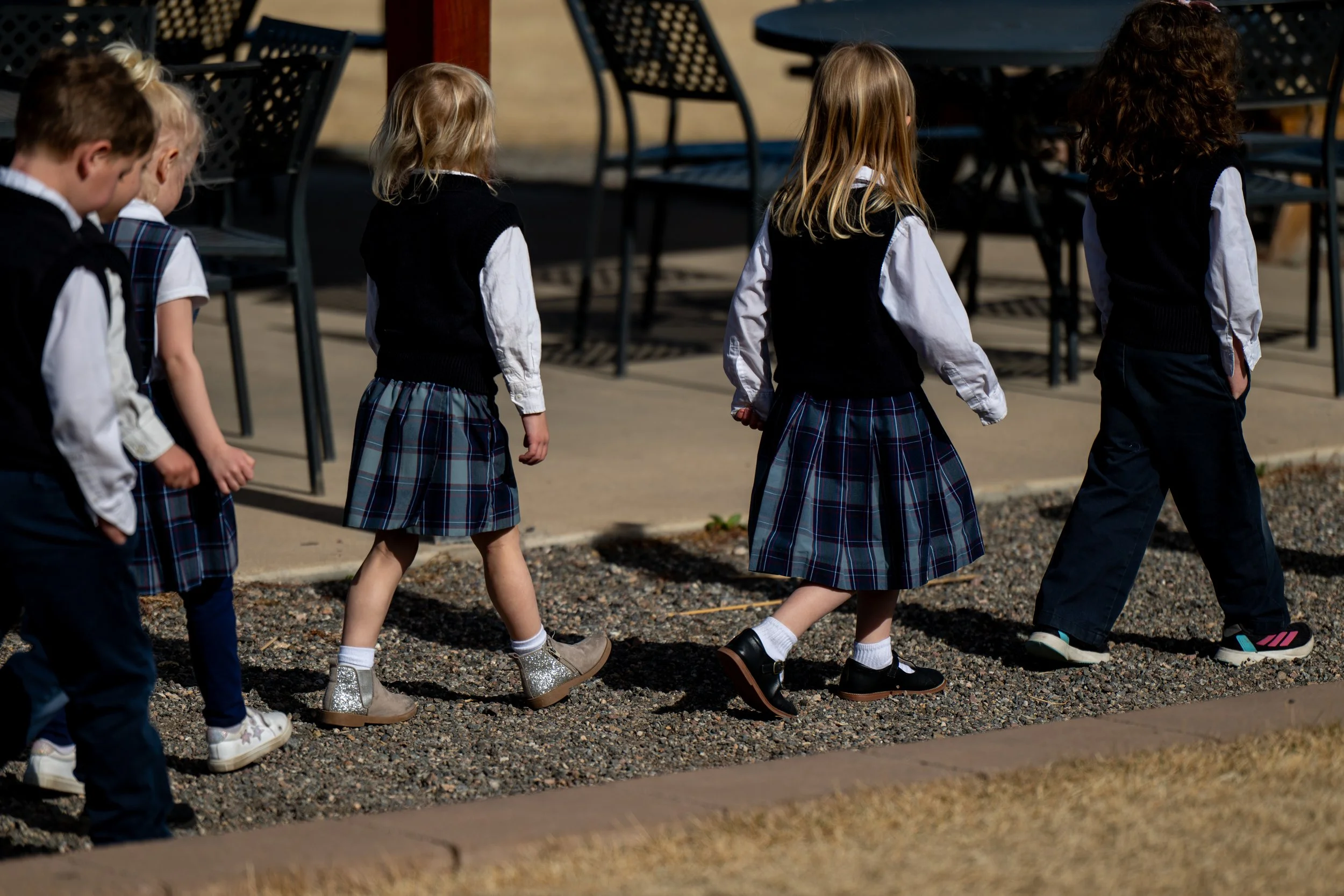 A group of children walking in a line outdoors, wearing school uniforms with plaid skirts and dark vests, with chairs and tables visible in the background.