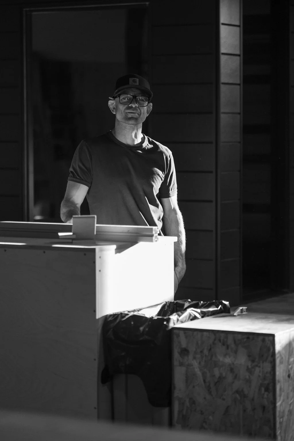 A man with glasses and a baseball cap standing next to a table inside a building with wooden paneling, in black-and-white photograph.