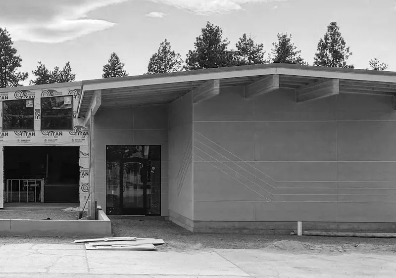 Front of a modern building under construction, with a flat roof, a large window, and a door, surrounded by trees and construction materials on the ground.
