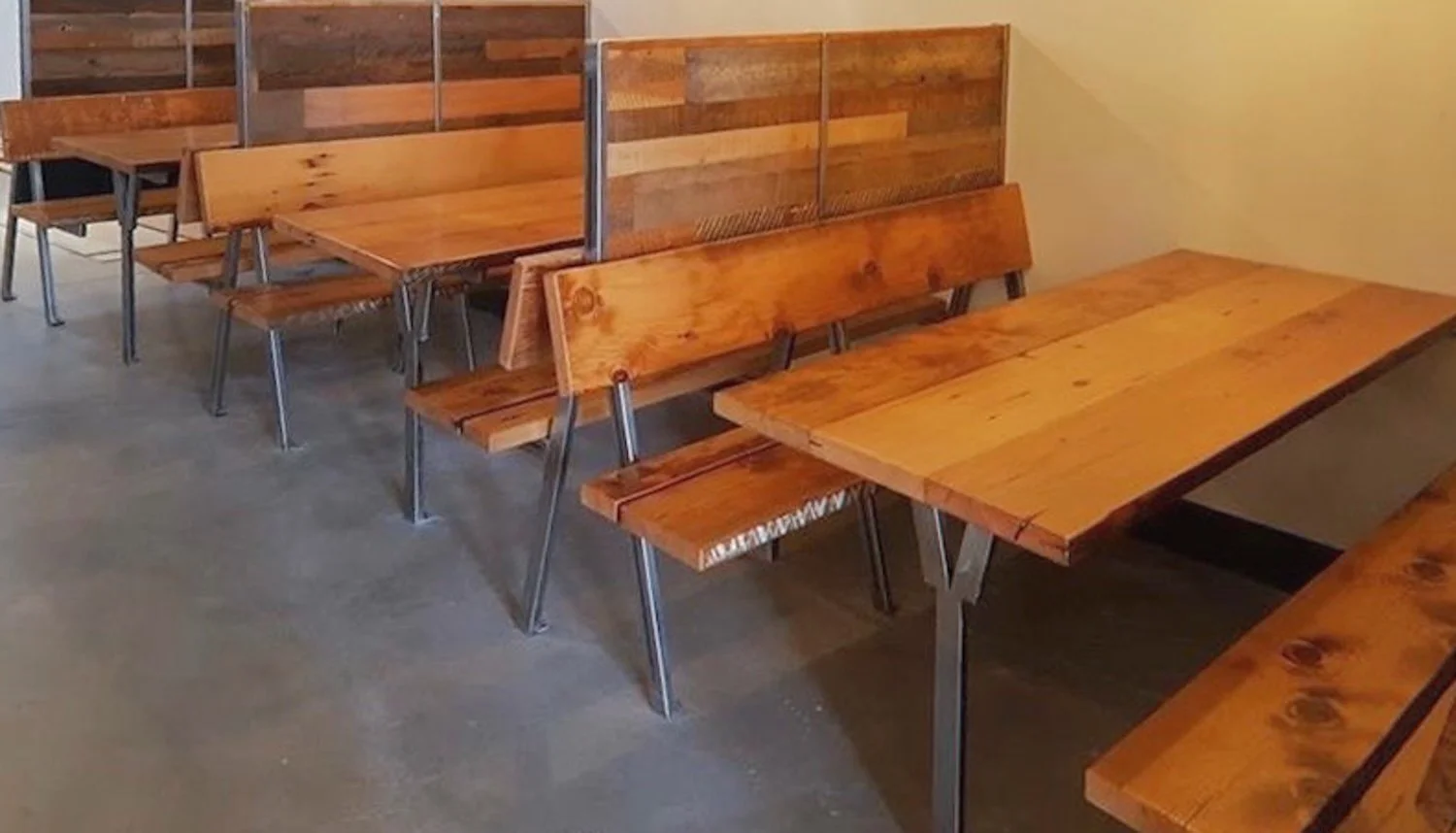 Wooden tables and benches in a dining area, with partition screens on some tables, against a plain yellow wall.