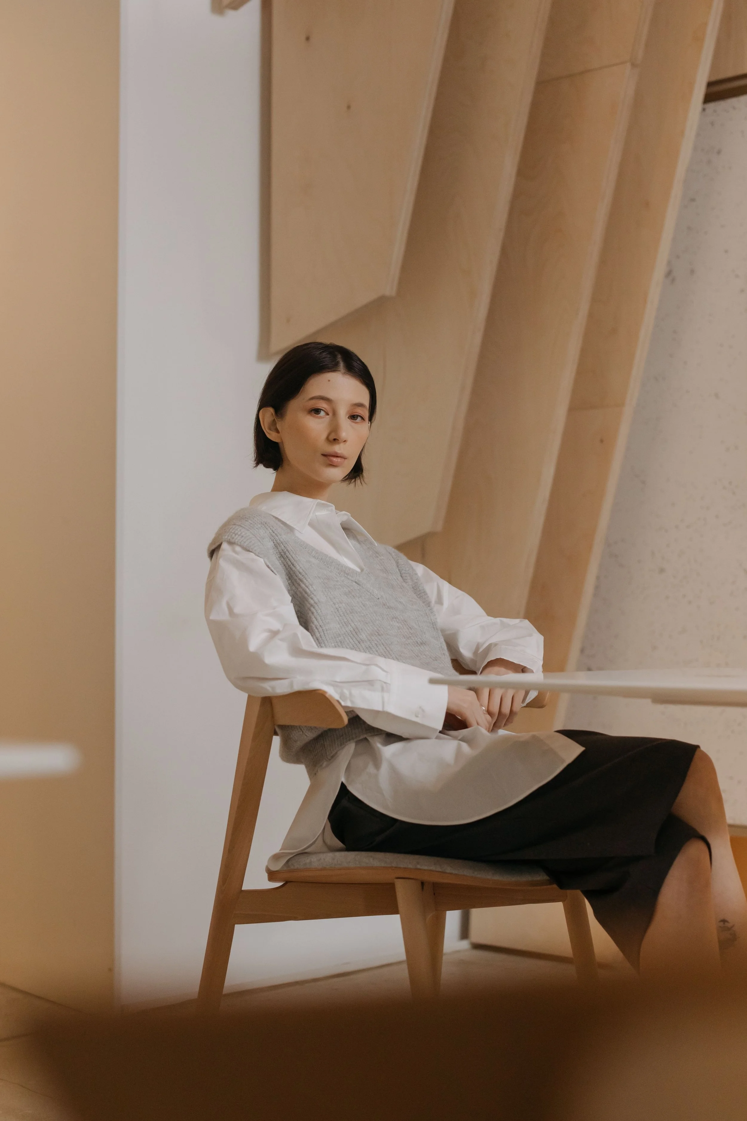 A young woman with short dark hair wearing a white shirt, grey vest, and black skirt, sitting on a wooden chair in a room with sloped wooden ceilings and light-colored walls.