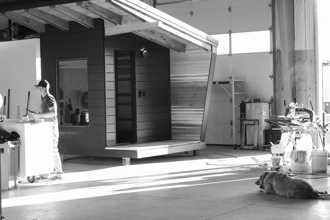 A person working inside a large garage or workshop with a partially built tiny house or small structure. The tiny house is on stilts with a front porch, and there are tools, boxes, and a dog lying on the floor.