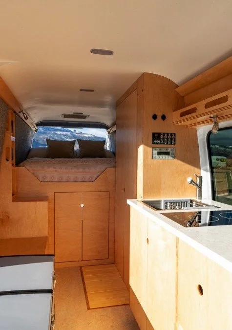 Interior of a camper or RV showing a small kitchen area with a sink, stove, and wooden cabinetry, and a sleeping area with a bed and pillows at the back. There's a window with a view outside.