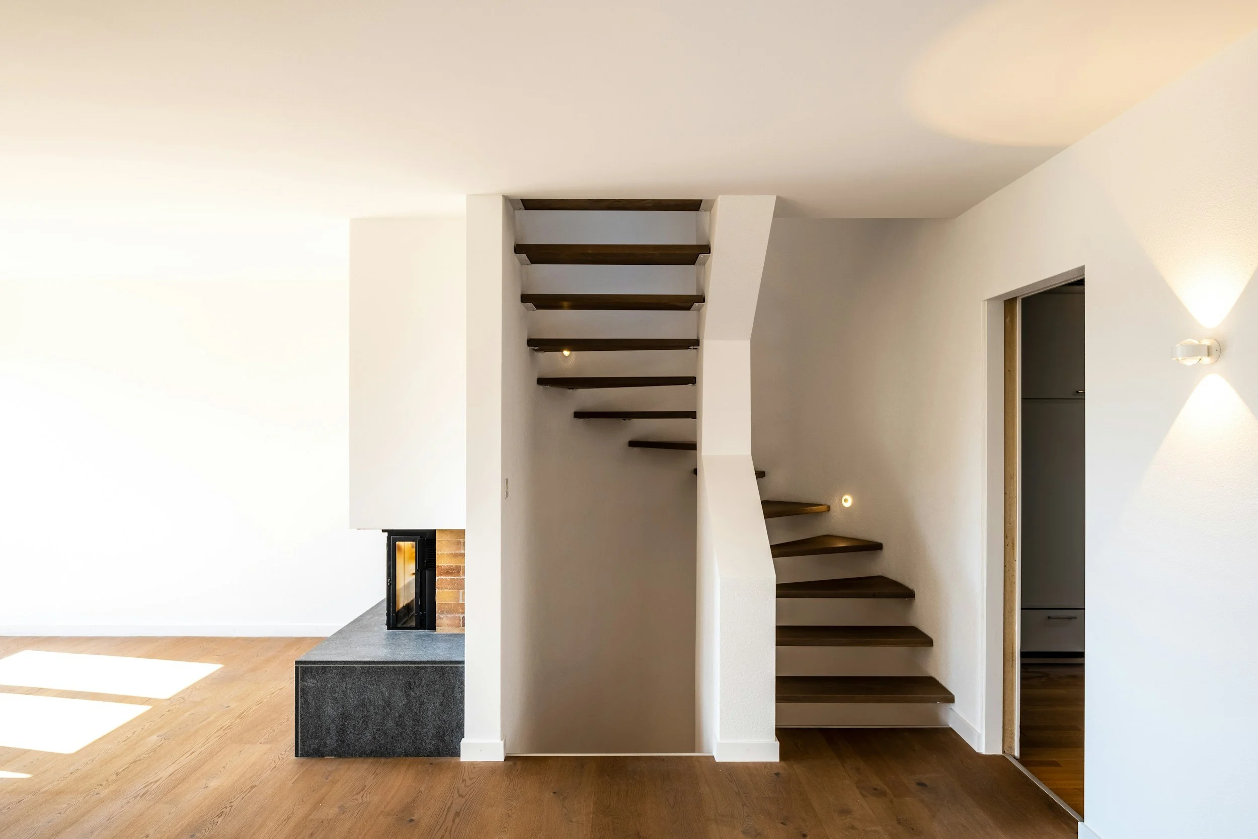 Interior view of a modern home with a staircase featuring wooden steps and a white wall, a fireplace with a brick and black frame, and hardwood flooring.