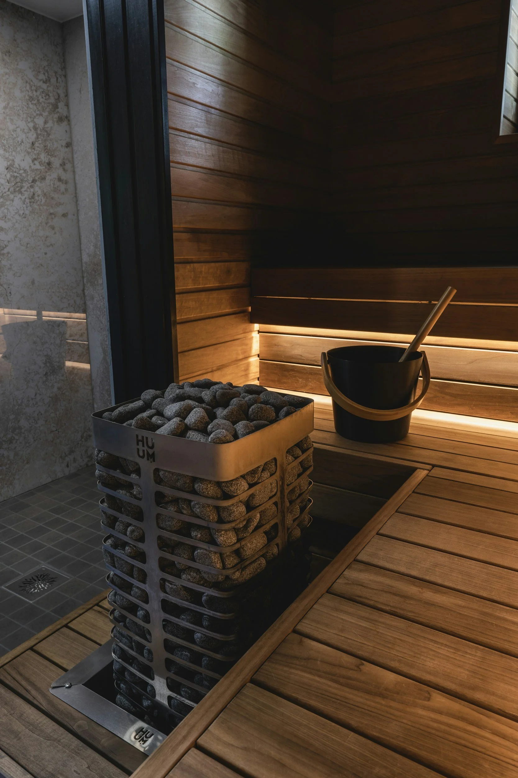 Indoor sauna or steam room with wooden walls and a wooden bench. A black bucket with a wooden ladle next to a basket filled with rocks or stones on a tiled floor.