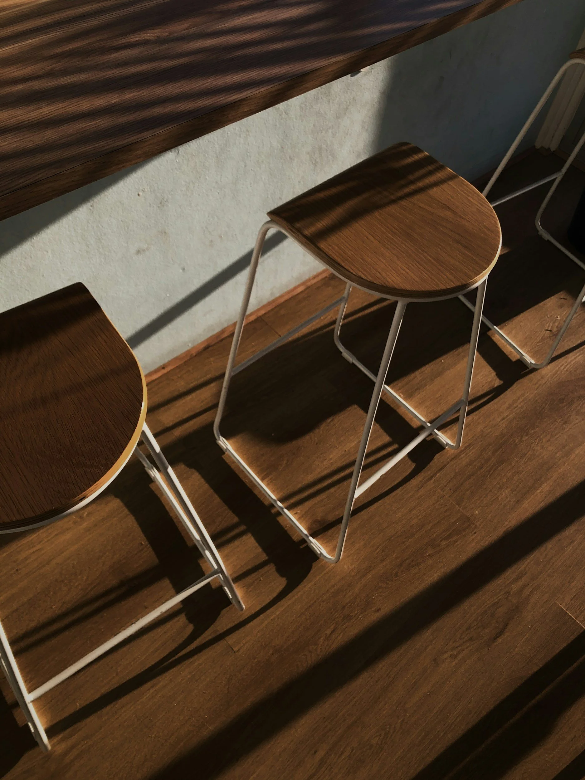 Wooden barstools with white metal legs against a textured wall, casting shadows on a wooden floor.