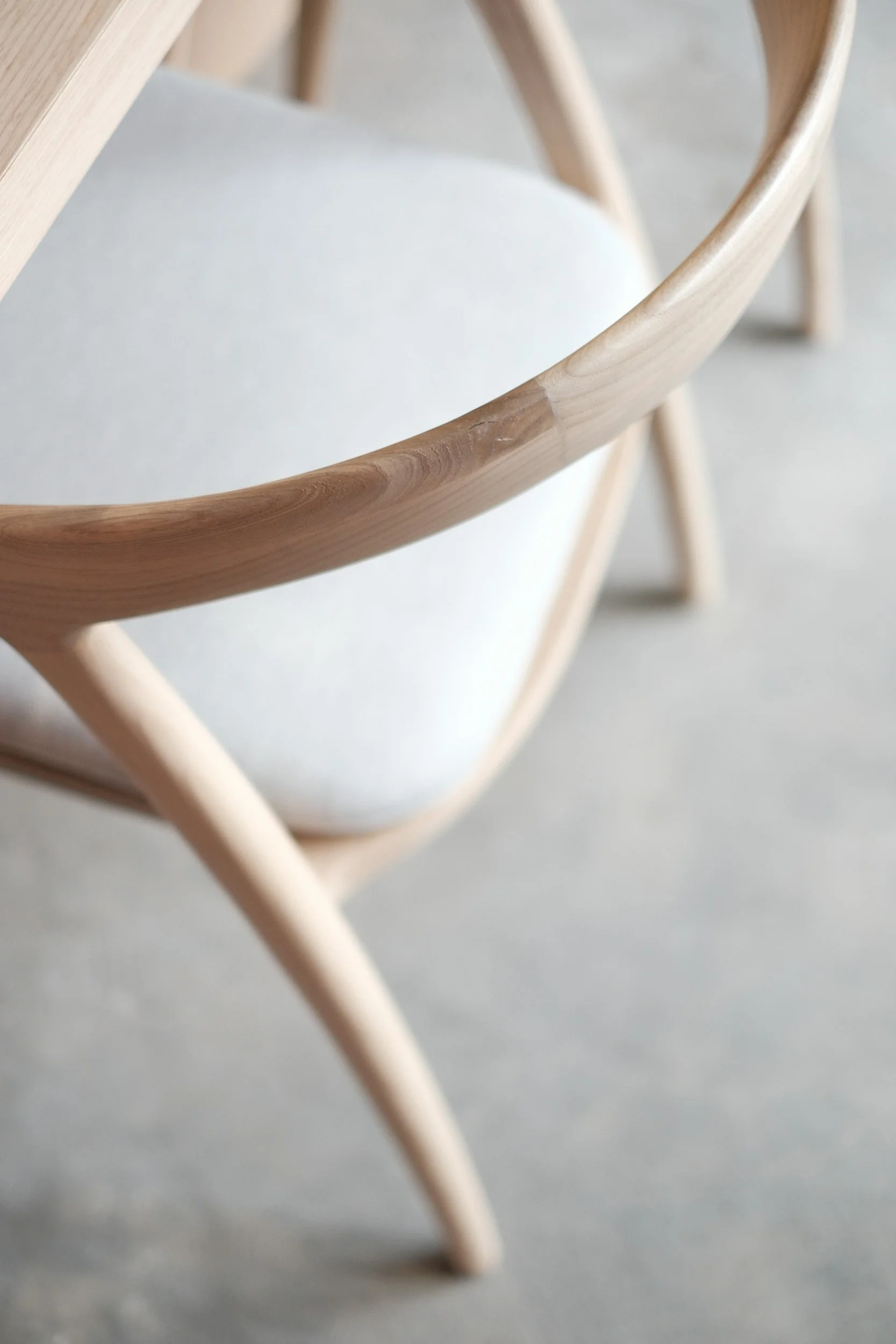 Close-up of a wooden chair with a curved wooden backrest and white upholstered seat, seen from an angle on a light-colored floor.