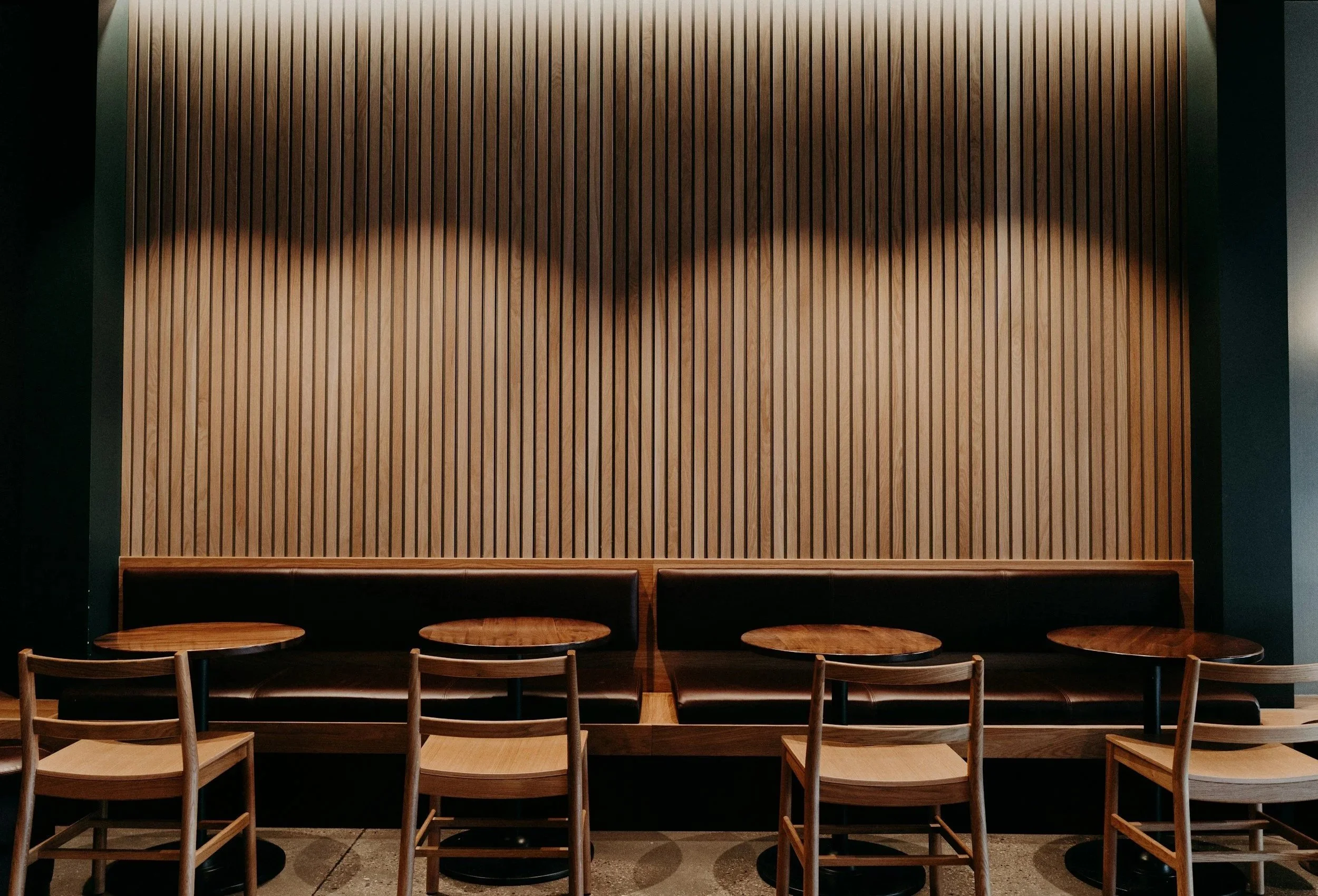 Interior view of a dining area with wooden chairs and tables lined in front of a large wooden panel wall.