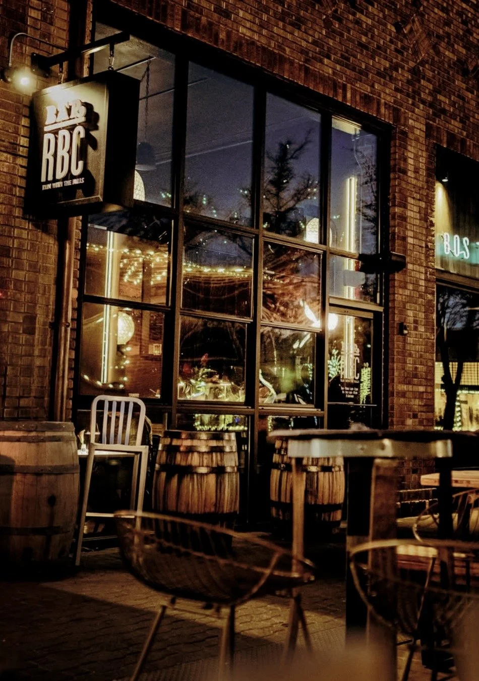 Nighttime exterior view of a restaurant or pub with large glass windows, brick walls, wine barrels, outdoor tables and chairs, and warm interior lighting.