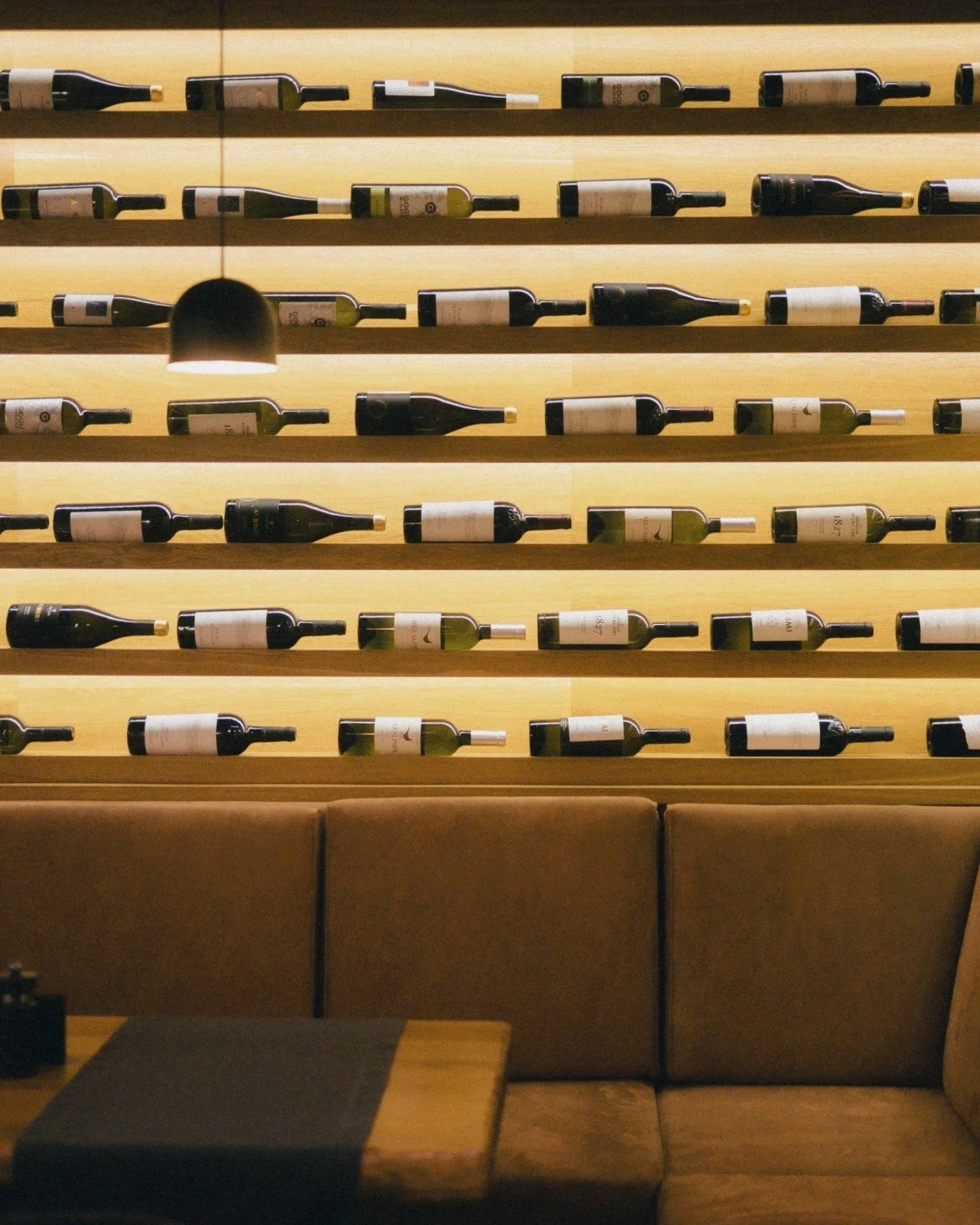 Wall-mounted wine bottles display with horizontal shelves, backlit with warm lighting, in a wine bar or restaurant setting.