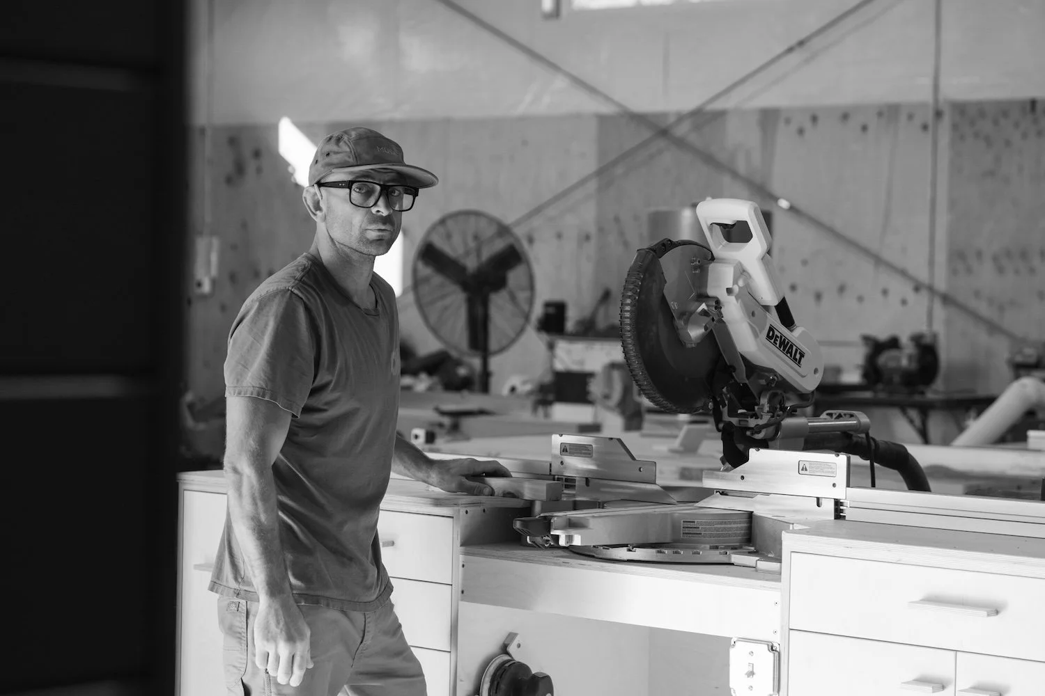 A man in a baseball cap, glasses, and T-shirt using a DeWalt miter saw in a woodworking workshop.
