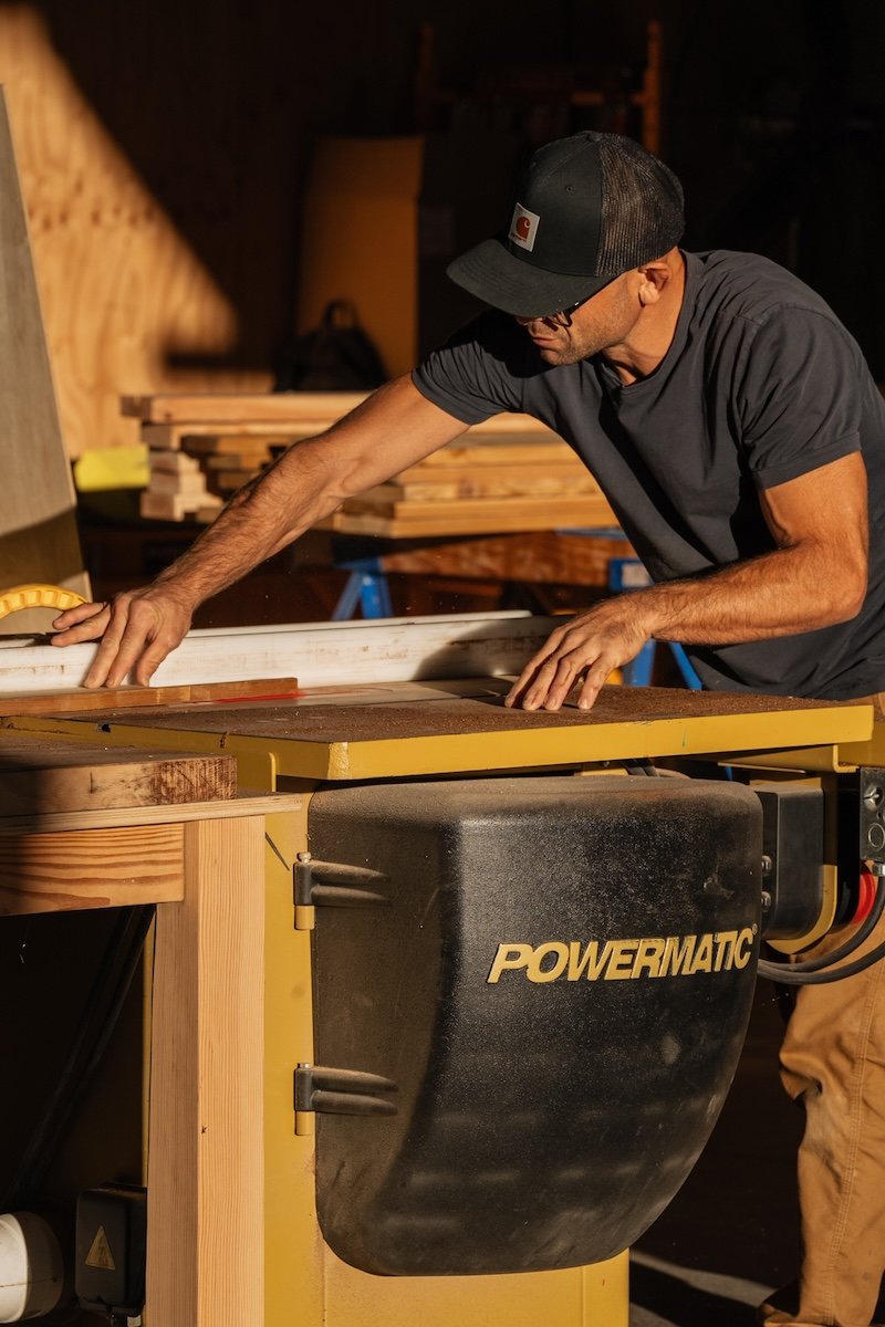 A man wearing a black cap and glasses is working in a woodworking shop, using a Powermatic table saw to cut a piece of wood.