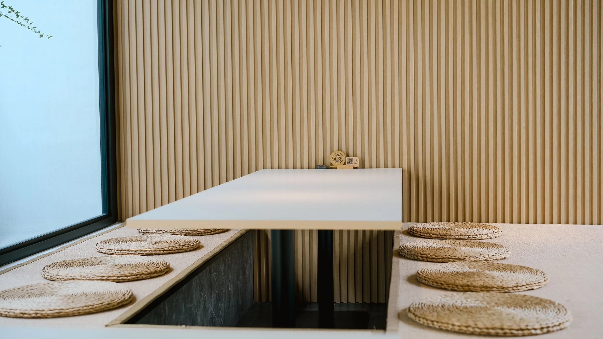 Minimalist dining or meeting area with a white table, woven seat cushions around a built-in bench, a wooden slat wall, a large window on the left, and a small sign on the table.