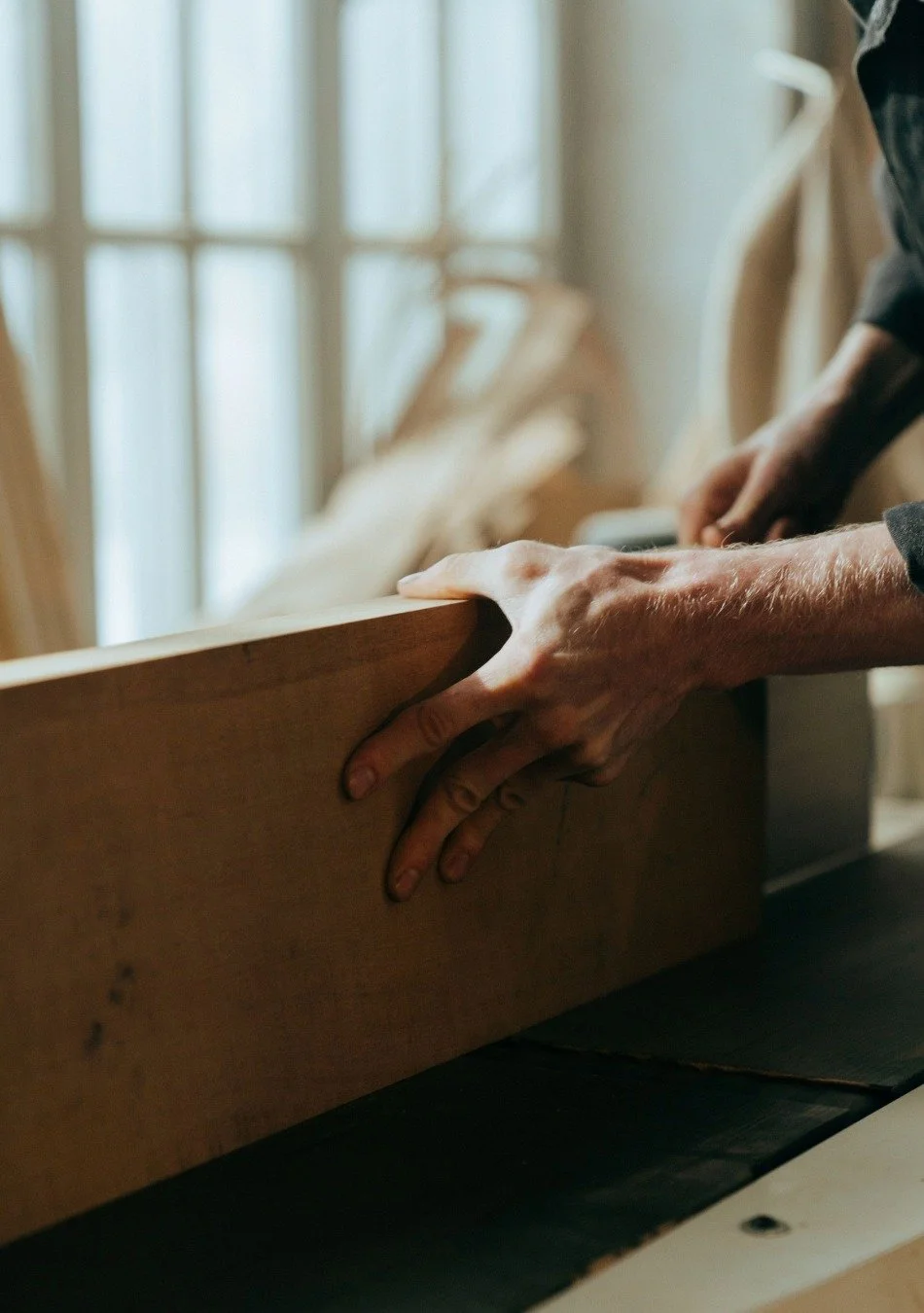 Close-up of person working with wood, shaping a wooden board with hands, sunlight coming through a window.