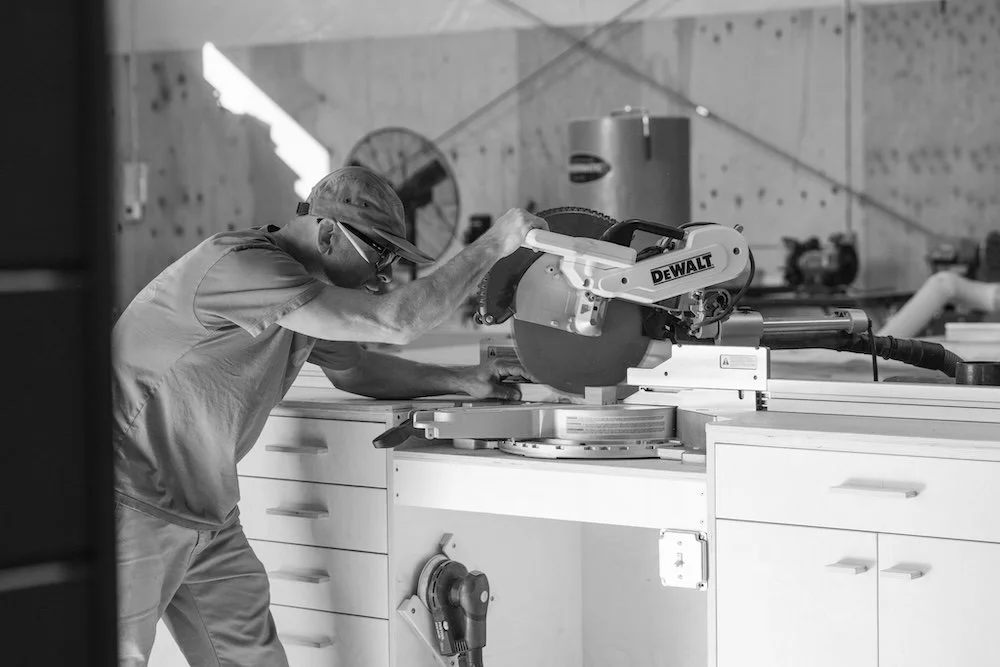 A man wearing safety glasses and a cap operating a large DEWALT miter saw in a workshop or construction site.