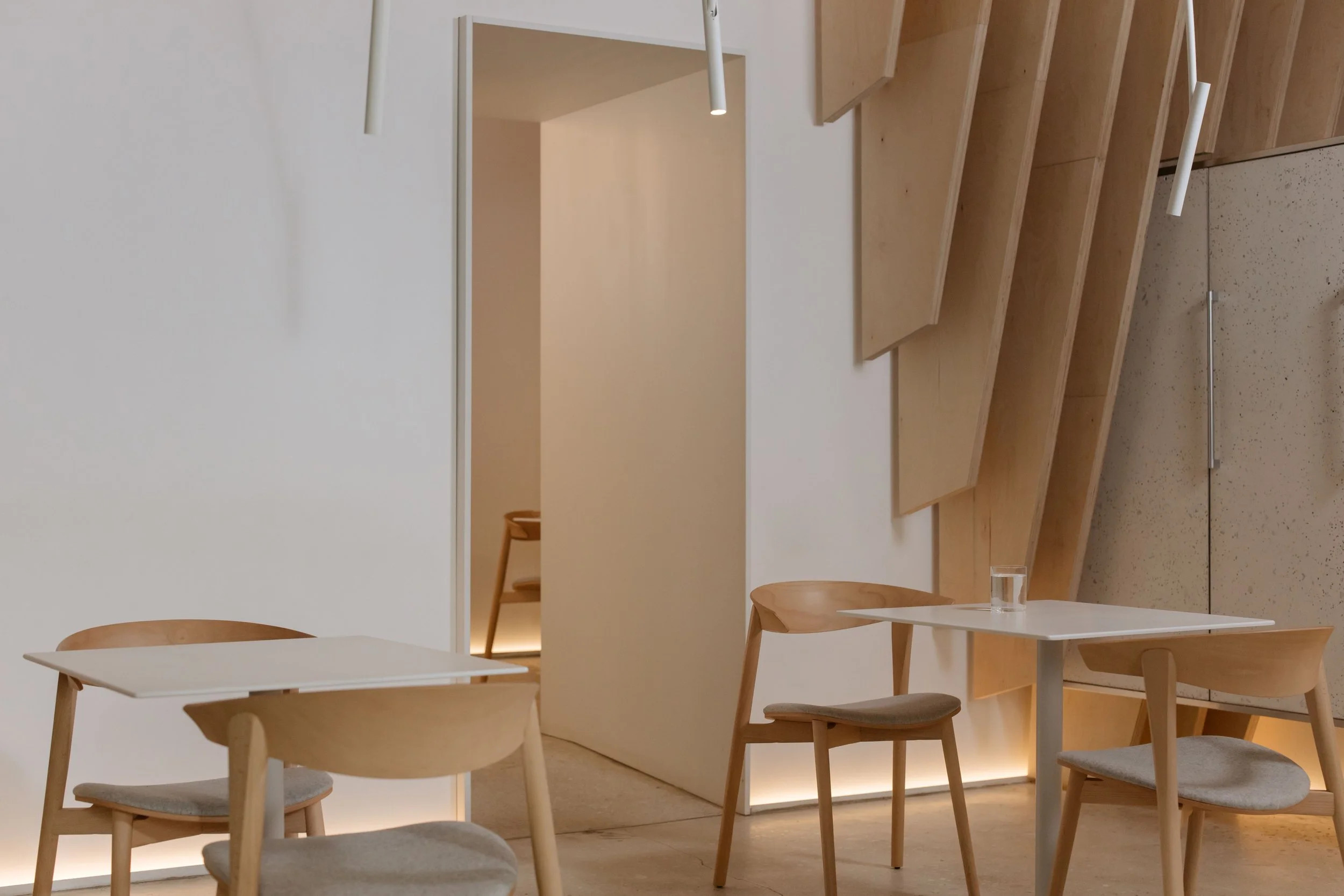 Interior of a minimalist, modern dining space with light wood furniture, white walls, and subtle lighting.