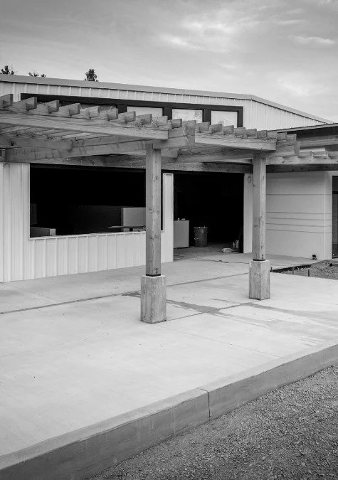 Black and white photo of a carport with wooden beams supporting a roof, attached to a building with siding. The area is paved with concrete, and there is a curb at the edge.