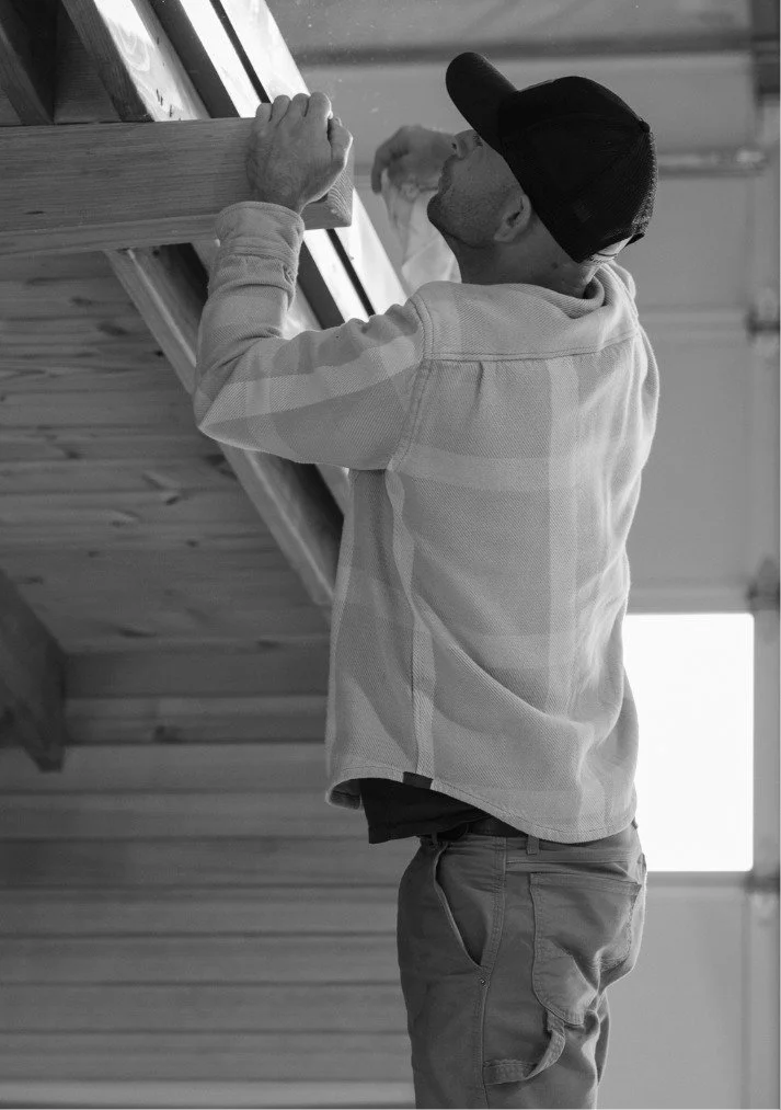 A man wearing a baseball cap and casual clothing working on or inspecting a wooden structure indoors.