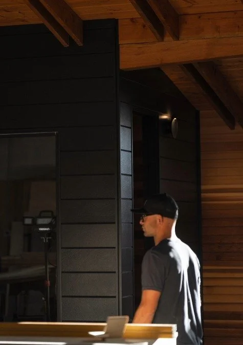 Side profile of a man wearing a black baseball cap and gray t-shirt standing indoors near a black wall and wooden ceiling.