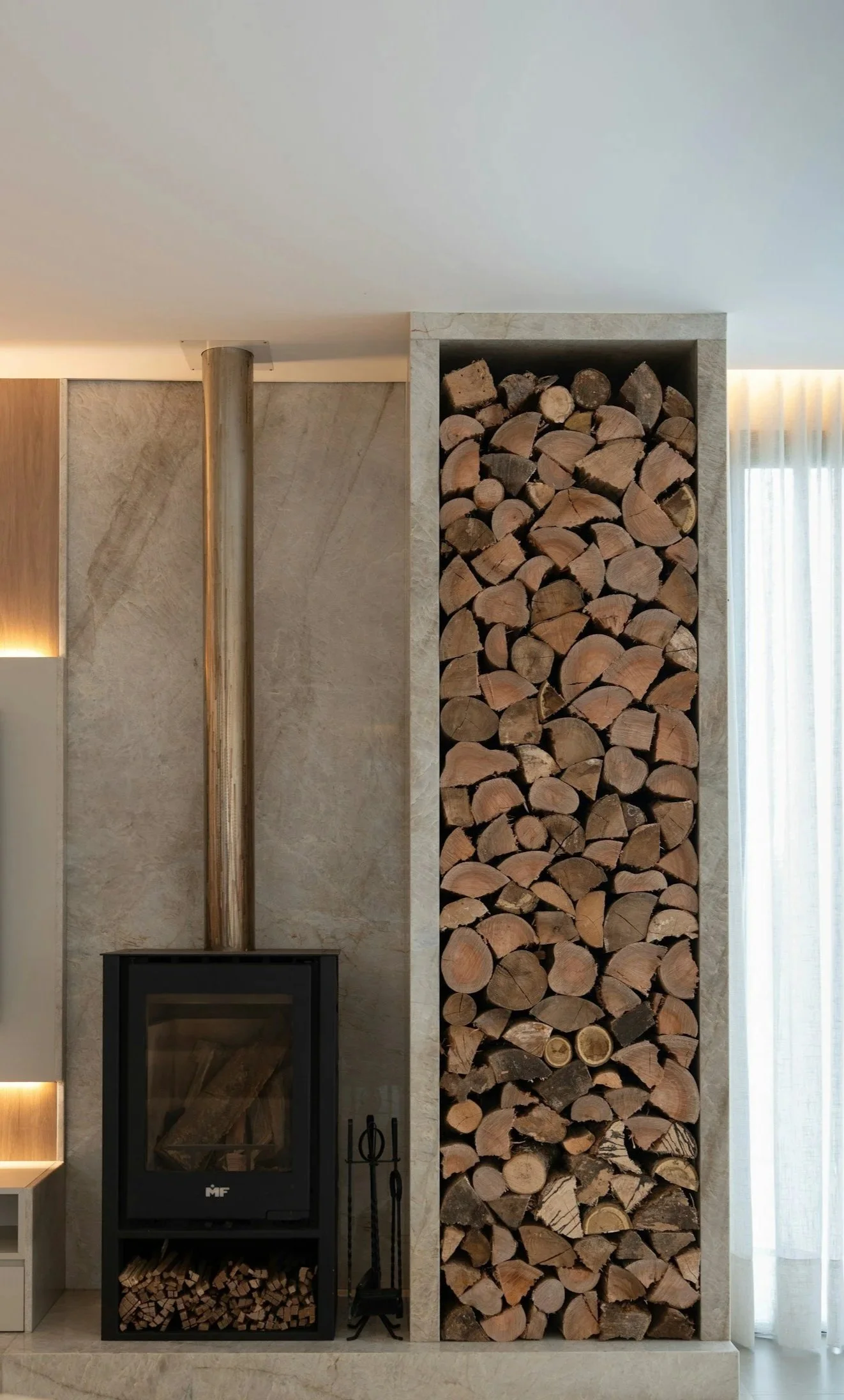 Wood pile stacked vertically next to a marble wall, with a corner of a fireplace and a metal chimney pipe visible above, in a modern living space.