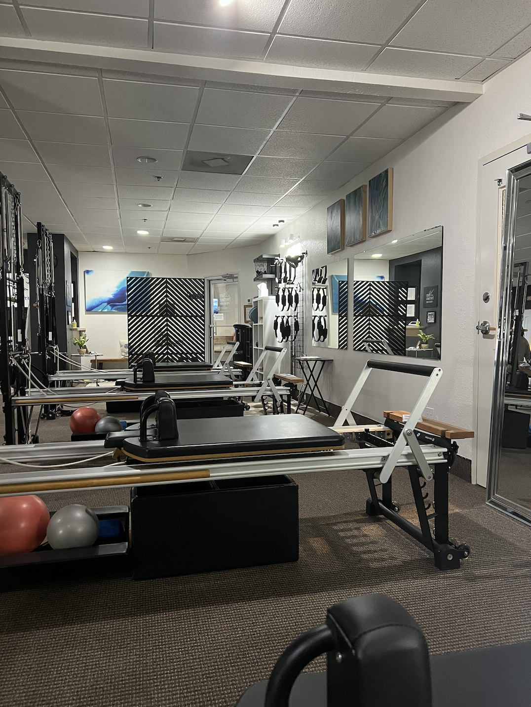 Pilates or physical therapy studio with reformer machines, exercise balls, wall-mounted resistance bands, mirrors, and wall art.