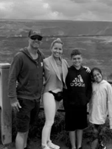 A family of four posing outdoors near a body of water, with a fence and landscape in the background. The group includes an adult male, an adult female, and two children, a boy and a girl.
