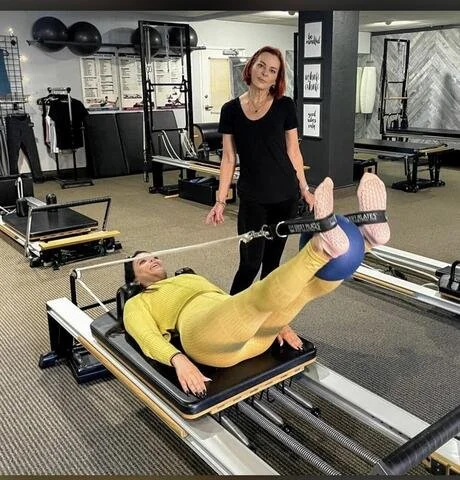 Woman exercising on a Pilates reformer machine in a gym, while another woman stands nearby observing.