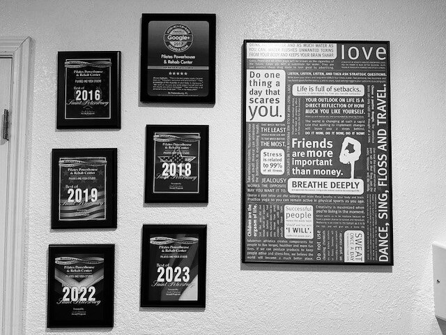 Wall display of framed plaques from 2016, 2018, 2019, 2022, 2023, and 2024, alongside a collage of motivational quotes and the word "love" in an office or studio setting.